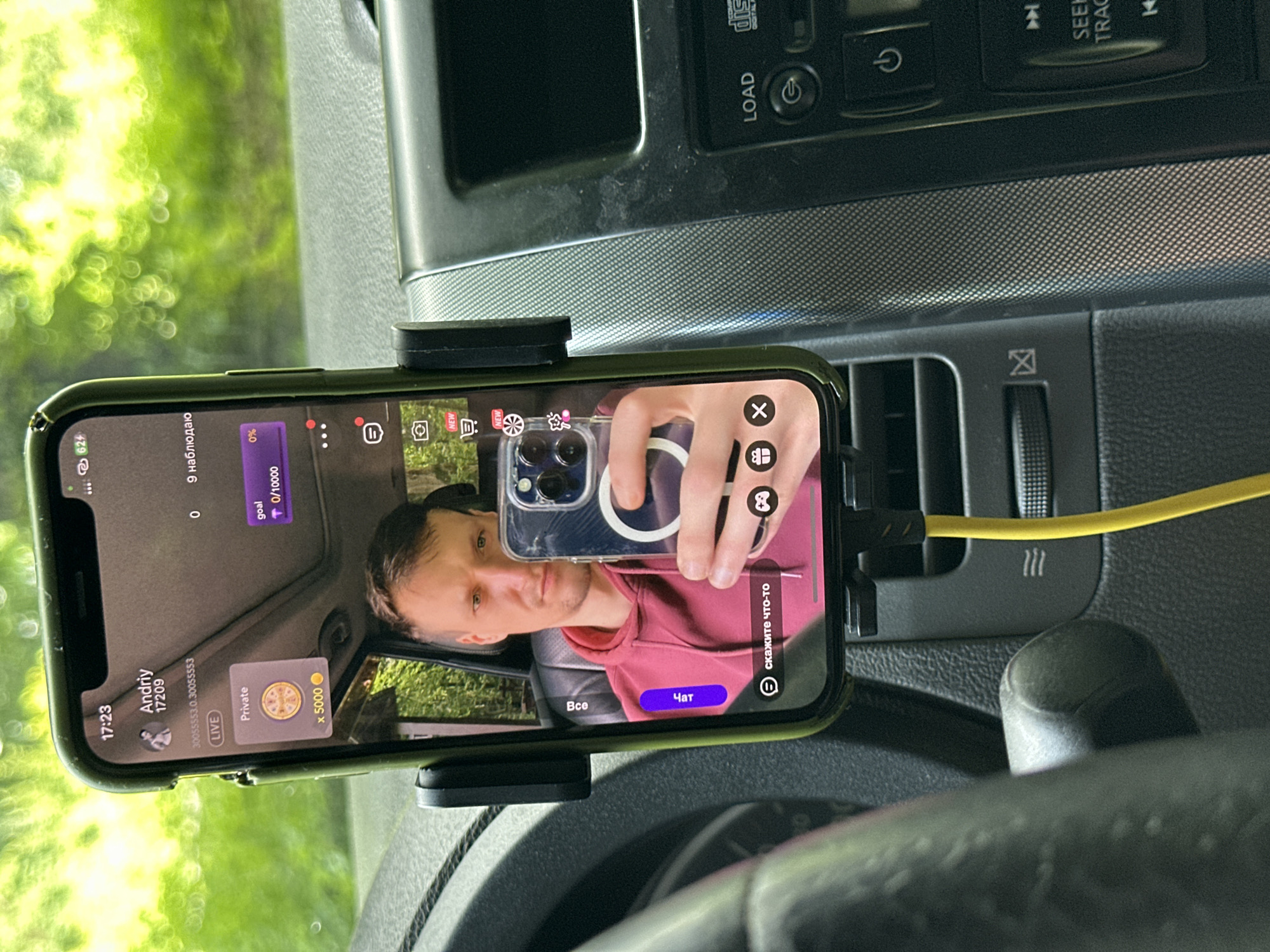 In the city of Shatava, Ukraine, a person is inside a vehicle, holding up a smartphone. The phone's screen displays a live stream interface. A man, identified as Andriiy, is visible in the reflection on the phone's screen, appearing to be the streamer. The phone is mounted on a car dashboard holder, connected to a yellow charging cable. The dashboard features various car controls and a car stereo system. The background seen through the car windows is lush green foliage, indicating an outdoor setting.