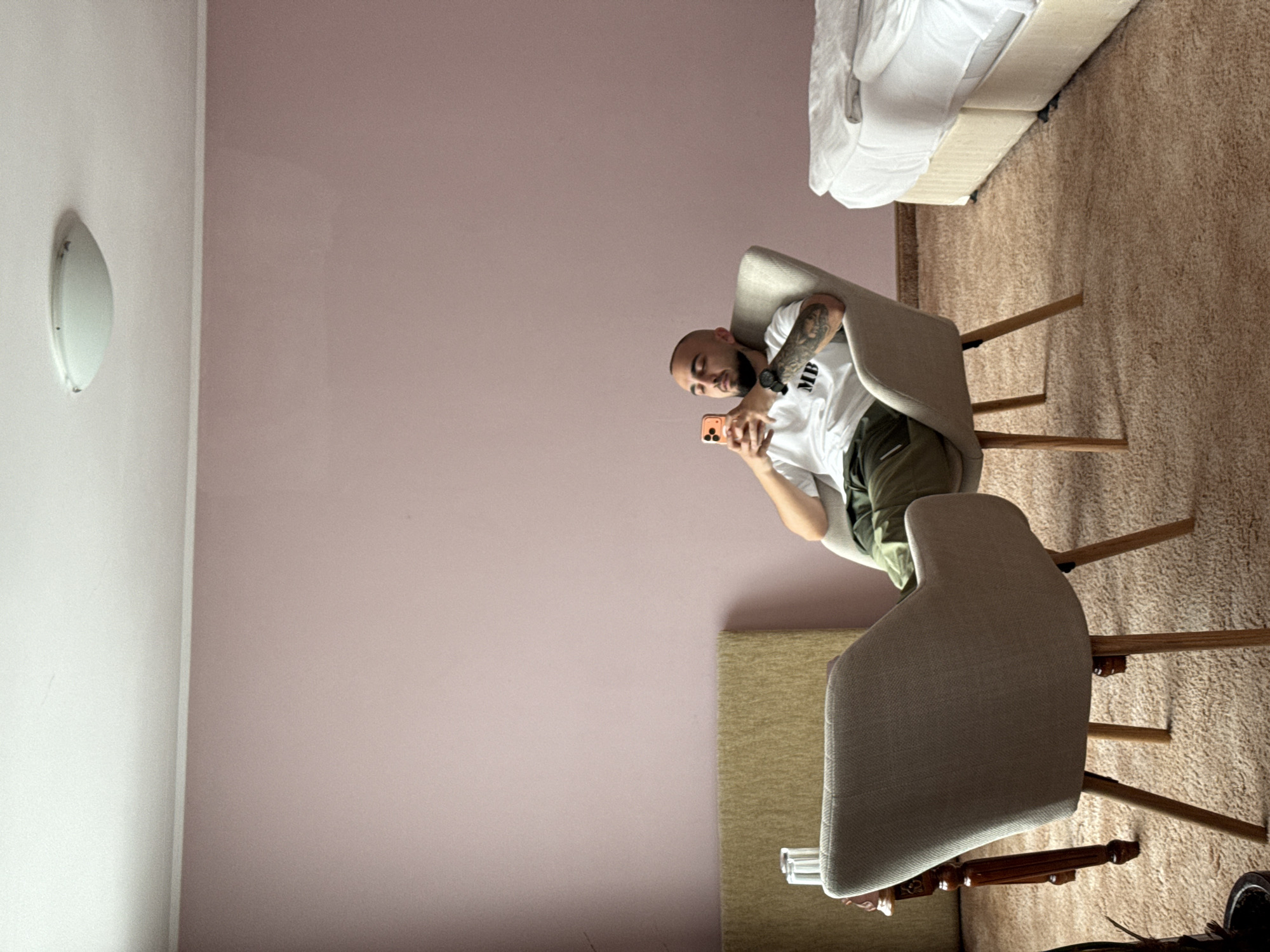 A bald man with tattoos on his arm is sitting in a chair and looking at his phone. The room has light pink walls, beige carpet, and a white lamp on the wall. There are two chairs in the room. The exact location in Veliko Tarnovo, Bulgaria cannot be determined from the image.