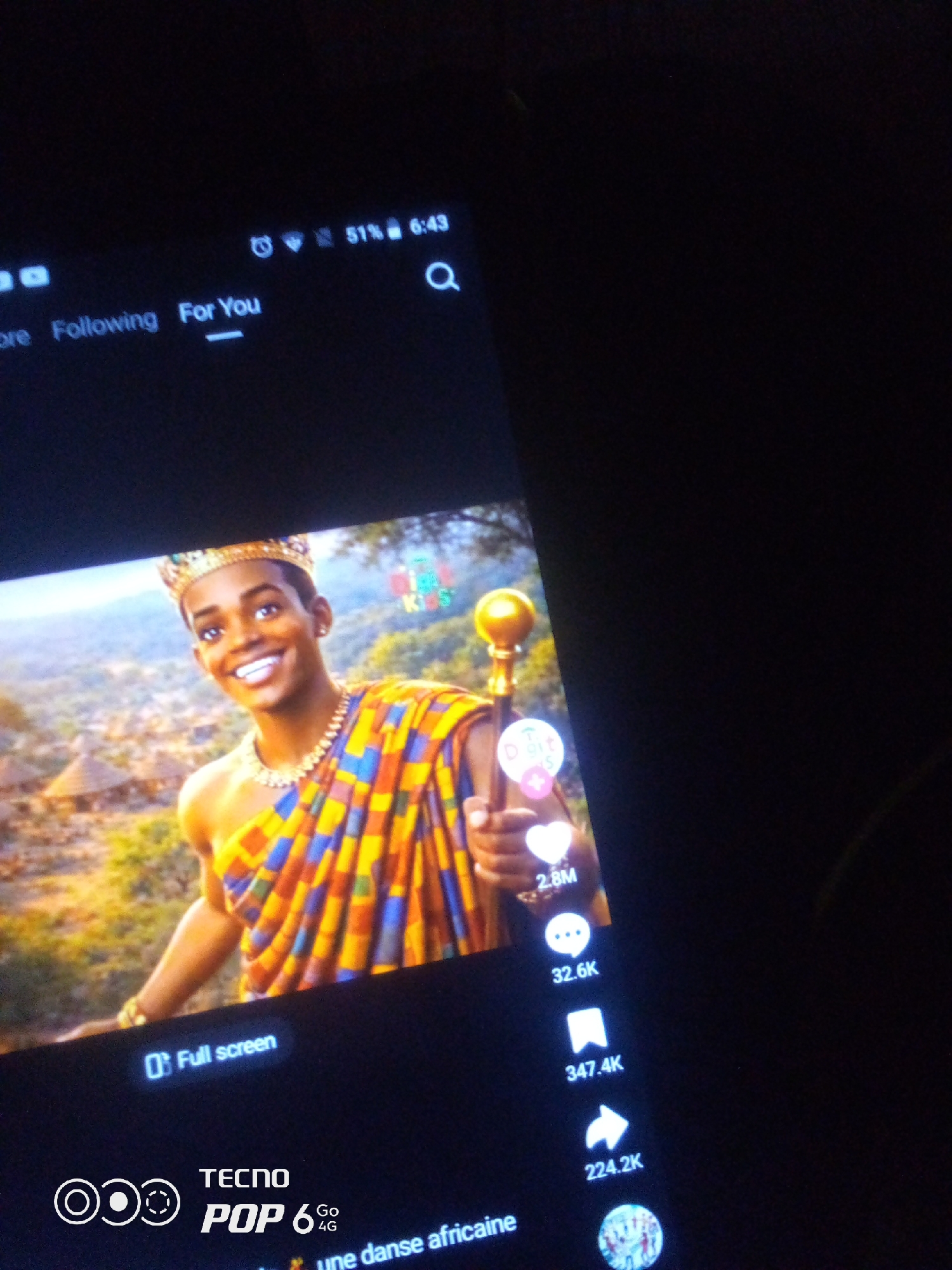 The image is a close-up shot of a smartphone screen displaying a TikTok video. The video features a smiling young man wearing a crown, a gold necklace, and a colorful, traditional African garment. He is holding a golden scepter in his right hand. In the background, there are huts and lush greenery, suggesting a rural or traditional setting.
On the screen, at the top left, "Following" and "For You" are visible, indicating the navigation options on the platform. The battery level shows 51% and the time is 6:43. On the right side of the screen, there are engagement metrics for the video: "2.8M" likes, "32.6K" comments, "347.4K" shares, and "224.2K" views. The bottom of the screen shows "Full screen" option and the branding "TECNO POP 6 Go 4G". A caption in French, "une danse africaine," is also partially visible. The device is held in a dark environment, implying the photo was taken in low light conditions. The image does not contain any information related to Howayi, Nigeria.