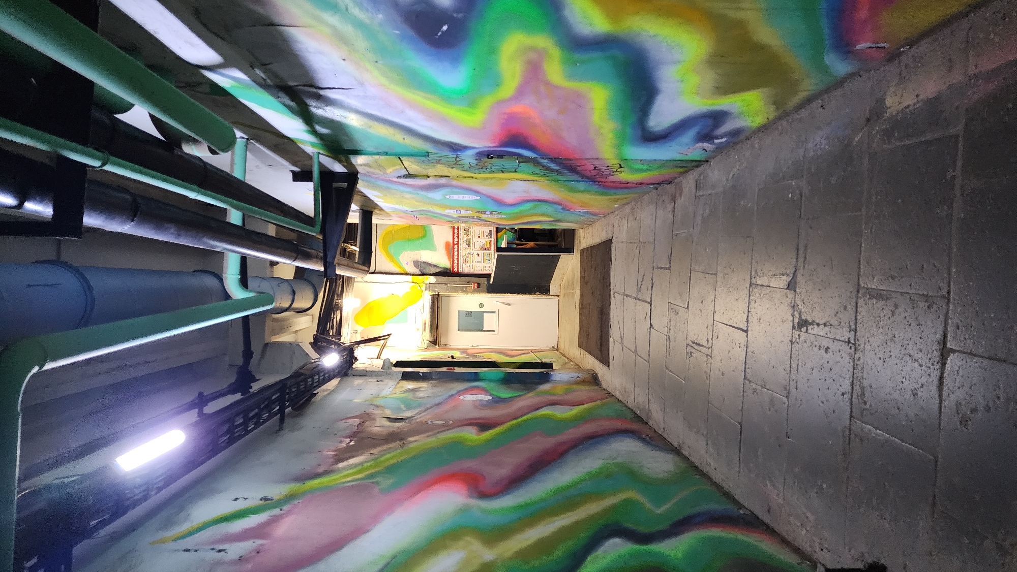 The image depicts an urban alleyway in Moscow, Russia. The alley's walls and ceiling are covered in vibrant graffiti art, featuring a mix of abstract shapes, colors, and patterns. Utility pipes run along one side of the alley, and a bright light fixture illuminates the area. At the end of the alley, a white door with a window is visible. The floor is paved with dark grey rectangular tiles. The overall environment appears somewhat gritty and neglected, typical of urban spaces. There are no people visible in the frame.