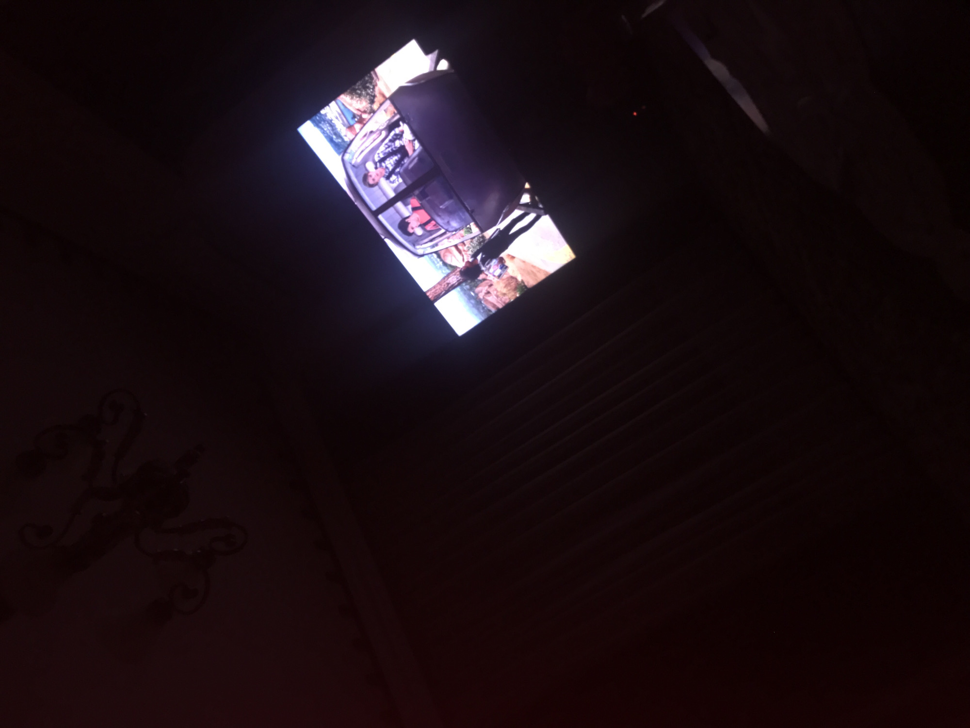 The image shows a dimly lit room in Yerevan, Armenia, with a television screen displaying a scene of people on a boat in a tropical setting. To the left of the frame, there is a decorative wall sconce. The room appears to have striped wall paneling.