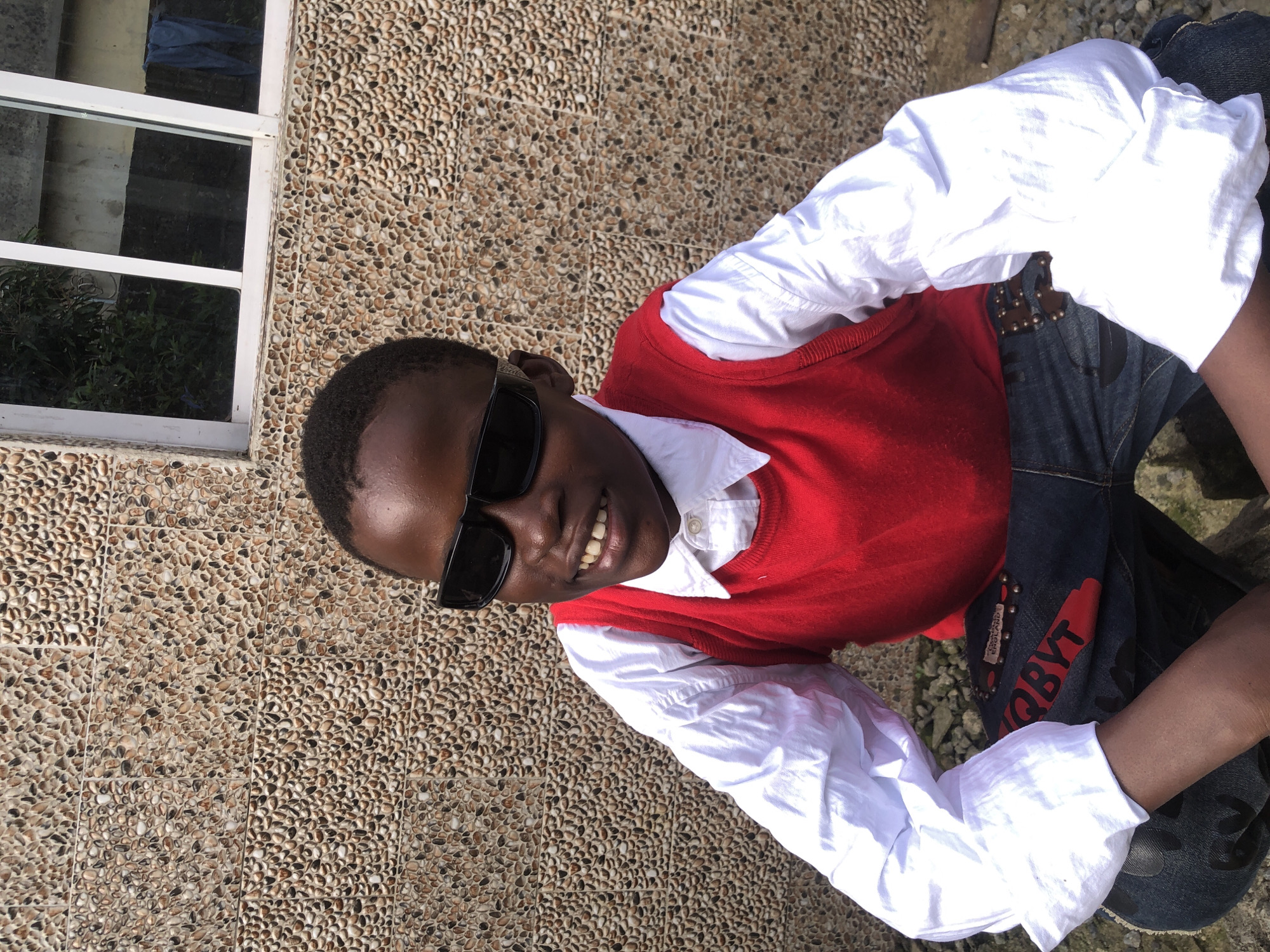 A young boy wearing sunglasses, a white collared shirt, a red vest, and blue jeans is crouched in front of a textured wall. The wall is made of small, irregularly shaped stones in various shades of brown, beige, and gray. To the right of the boy, a portion of a white-framed window with multiple panes is visible. Through the window, greenery and a light-colored building can be seen. The ground beneath the boy appears to be rough and uneven, with some small stones and debris. The scene is outdoors, likely in Ado, Nigeria, based on the provided context.