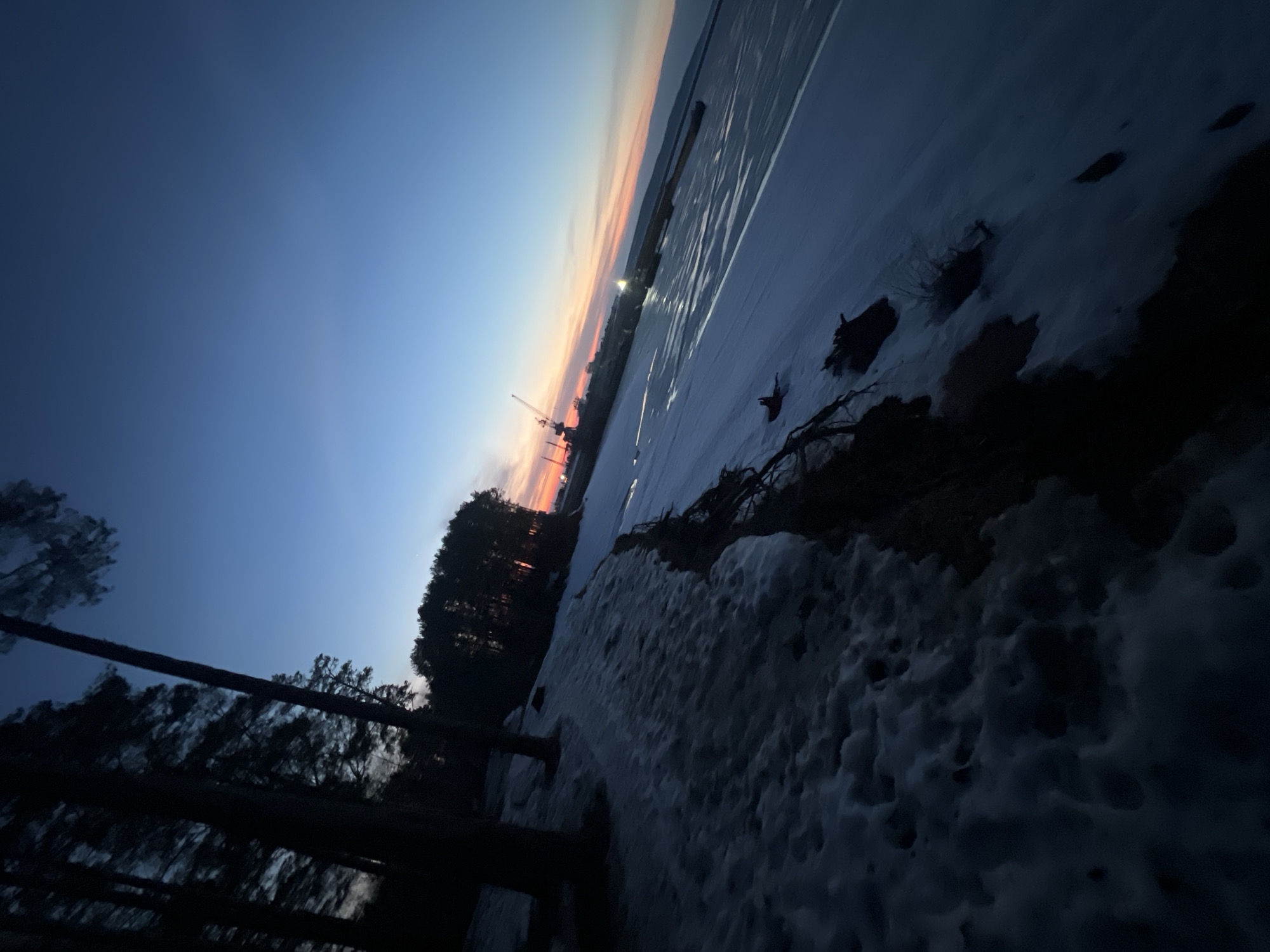 The image shows a snow-covered landscape at dusk in Chaikovsky, Russia. The scene includes a frozen body of water, likely the Kama River, with evidence of tracks on the ice. A treeline marks the edge of the water, and a crane and other industrial structures are visible in the background. Snow covers the foreground, with patches of exposed earth and vegetation. The sky displays a gradient of colors from blue to orange and red on the horizon, indicating either sunset or sunrise. There are no people visible in the frame.
