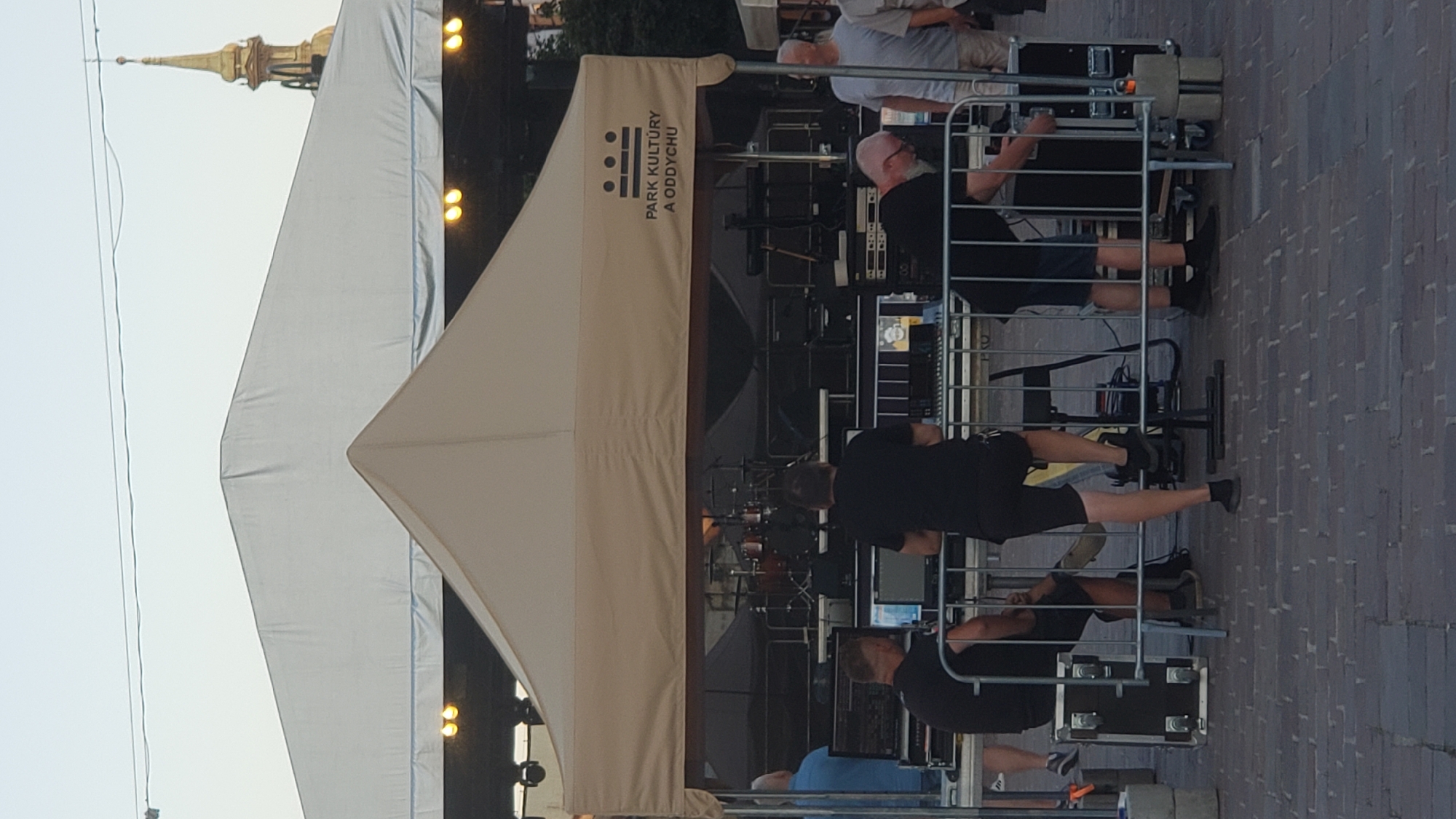 The image depicts an outdoor event setup, likely for a performance, in front of a large white tent. A smaller beige tent is positioned in front of the large tent, displaying a logo and text that reads "PARK KULTÚRY A ODDYCHU". This likely indicates the location is the Culture and Recreation Park in Prešov, Slovakia.
Several individuals are visible, appearing to be involved in the technical setup of the event. In the foreground, to the left, a person wearing a black t-shirt and shorts is seated and looking at a laptop. Next to them, another person in a black t-shirt and shorts stands behind a console with screens and equipment. To the right, a person with a shaved head, wearing a black t-shirt and shorts, is interacting with equipment. Further to the right, another person in a light-colored t-shirt and shorts stands observing.
Behind the beige tent, musical instruments, specifically a drum set, are visible, suggesting a band setup. The ground is paved with brick. The overall atmosphere suggests preparation for an event, with a focus on sound and stage equipment. The time of day appears to be late afternoon or early evening, given the lighting. In the background, a golden spire of a building is visible, possibly a church steeple, adding to the urban setting.