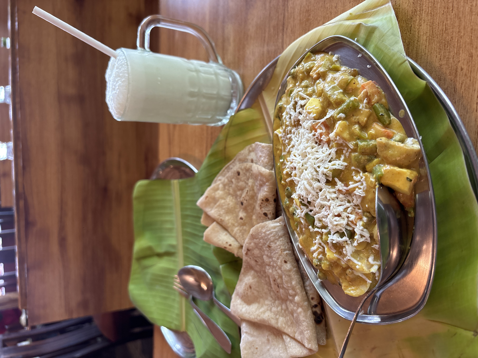 The image captures a meal presentation likely in Kamalapura, India, evidenced by the cuisine and style of serving. On a wooden surface, there's a layered serving arrangement. At the base is a metal tray layered with broad green banana leaves. On the right, a silver oblong dish contains a yellow, creamy vegetable curry topped with shredded white cheese. A spoon is inserted into the curry. To the left of the curry, stacks of chapati bread are neatly placed on the banana leaves. Beside the chapati, a spoon and a fork rest on the green leaves. In the background is a glass mug filled with a frothy white beverage, possibly lassi, and a white straw. The mug has a handle and sits atop the edge of the tray. The light suggests daytime. The environment appears to be a restaurant or eatery due to the presentation style. No people are visible in the image.