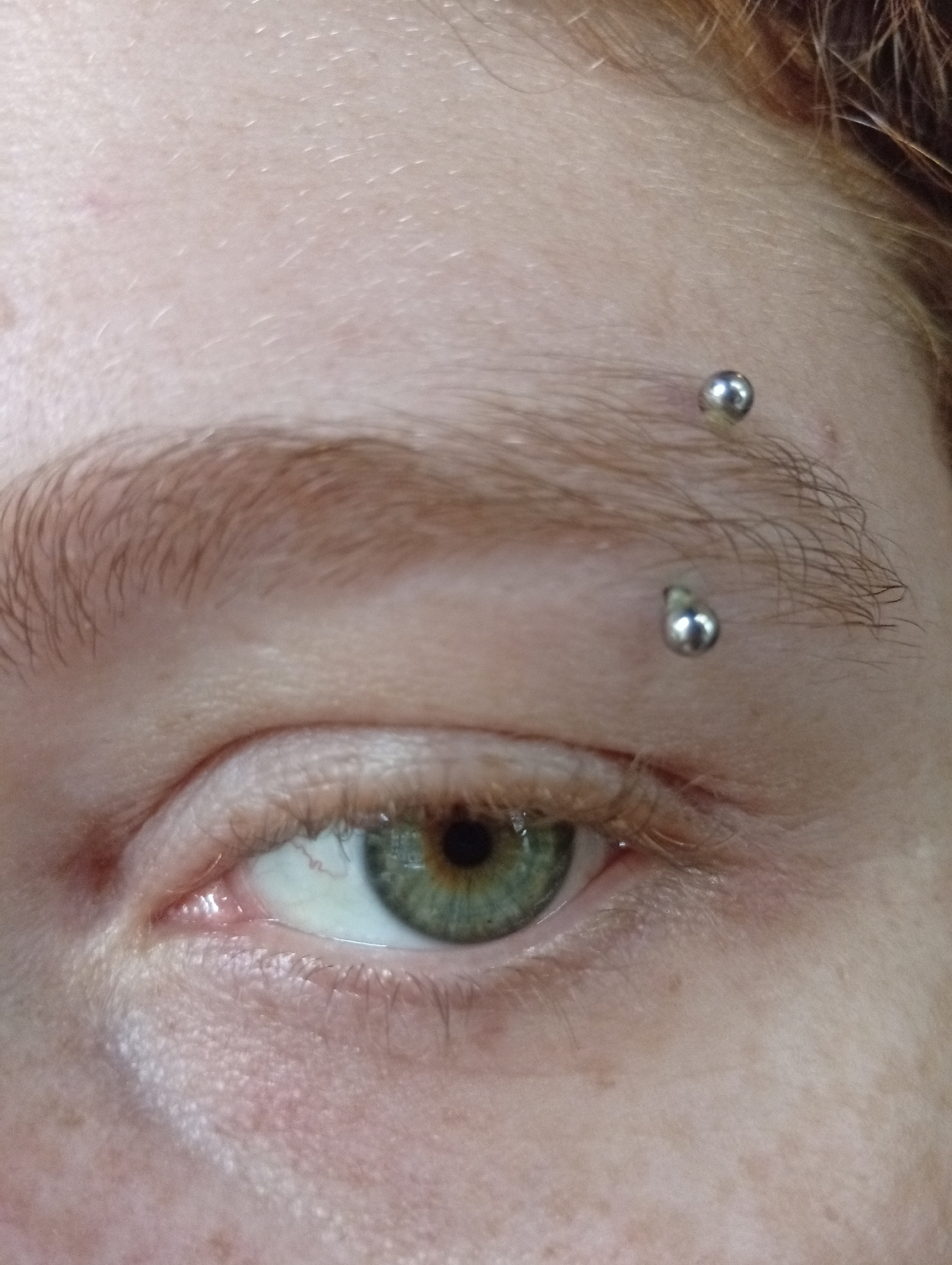 A close-up view of a person's left eye and eyebrow. The eye is green with a dark pupil. The eyebrow is light brown and pierced twice with what appears to be silver barbells. Skin tone is fair with freckles. No elements indicate a location in Buenos Aires, Argentina.