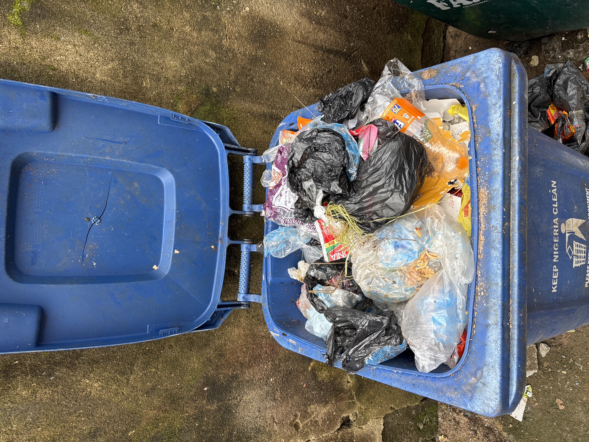 An overhead shot captures a blue plastic trash bin overflowing with refuse. The bin's lid is open, revealing a jumble of plastic bags, some containing visible trash. Among the debris are a torn orange plastic bottle and various wrappers. The exterior of the bin has the slogan "KEEP NIGERIA CLEAN" printed on it. The bin sits on a rough, dirty concrete surface. To the right of the blue bin, a sliver of a green bin is visible. The surrounding environment appears to be outdoors, possibly in an urban setting in Ihiagwa, Nigeria, given the visible text on the bin. The lighting suggests it is daytime.