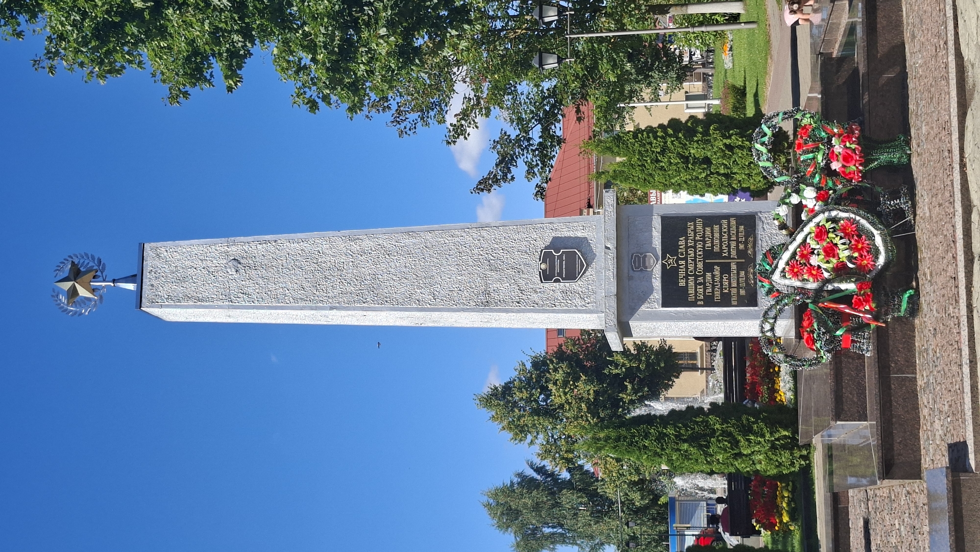 This is a photograph of a memorial monument located in the city of Pastavy, Belarus. The monument is an obelisk made of light-colored stone, topped with a golden star within a laurel wreath. The base of the obelisk features a dark plaque with inscriptions in Cyrillic, listing names and dates, and a shield emblem. Below the plaque, several large, circular wreaths with red and white flowers and green ribbons are placed. The monument is situated in a well-maintained public space with manicured evergreen trees, flower beds with colorful blossoms, and a fountain in the background. A lamppost with two lights is visible to the right, and a red-roofed building can be seen behind the trees. The sky is a clear blue with a few wisps of clouds. In the foreground, a person, appearing to be a child, is interacting with the base of the monument. The overall scene depicts a solemn and commemorative outdoor setting.
