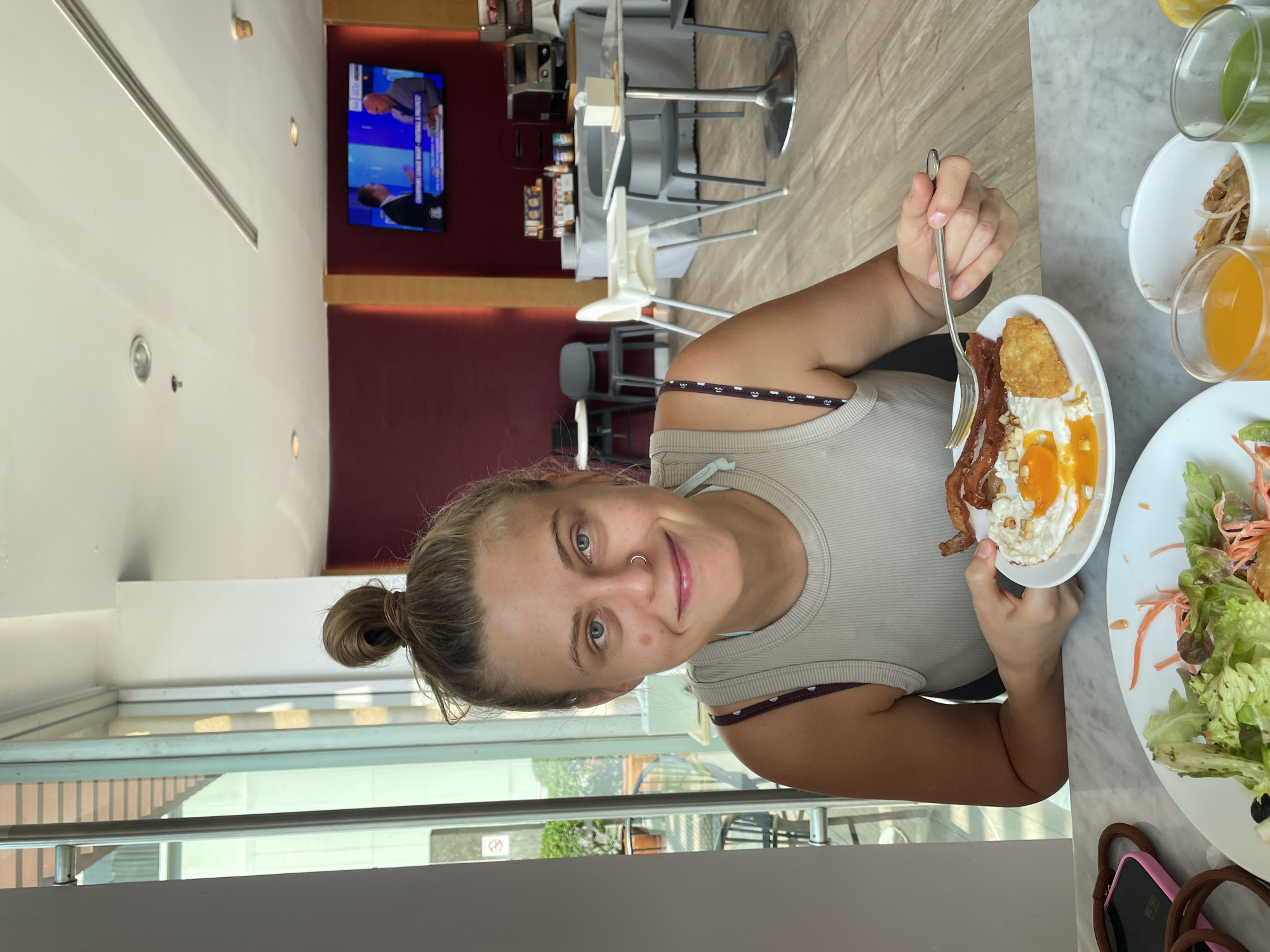 A young woman with blonde hair in a bun is seated at a table in what appears to be a hotel dining room in Bangkok, Thailand. She is holding a plate of breakfast food including bacon, eggs, and a hash brown. There is also a plate of salad and several glasses of juice on the table. A television showing a news program is visible on the wall in the background. The dining room has modern décor and is well lit. The woman is smiling and looking directly at the camera. Through the window on the left there are exterior buildings visible.
