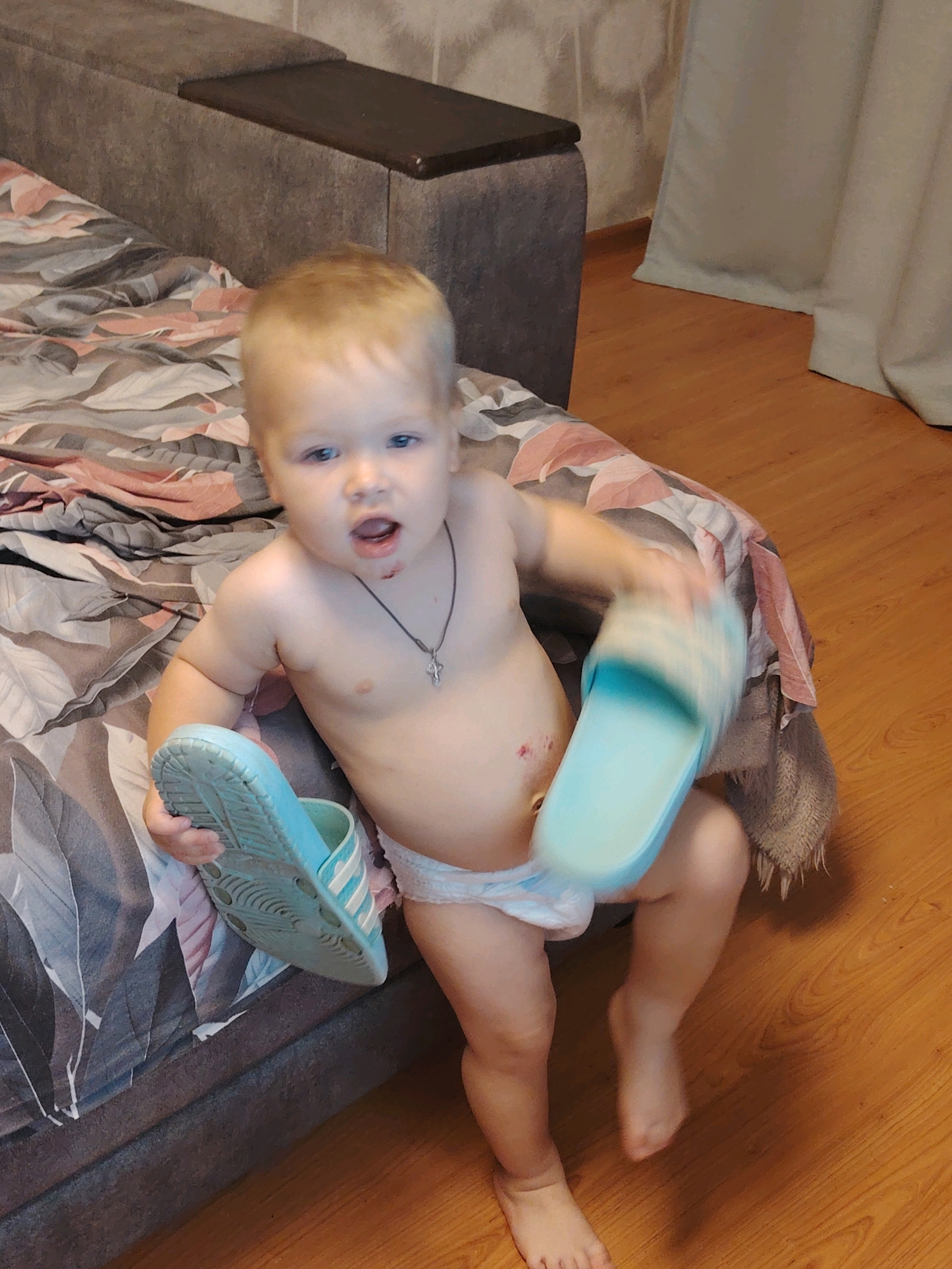 A blonde, fair-skinned baby wearing a diaper and a necklace is sitting on a bed with a patterned blanket. The baby is holding two light blue and white striped slides. The baby's mouth is open, and there appears to be a small mark on their chin. The bed is next to a dark grey headboard with a wooden top. In the background, there is a wooden floor and a light-colored curtain. The location is identified as being in the city of Chaikovsky, Russia.