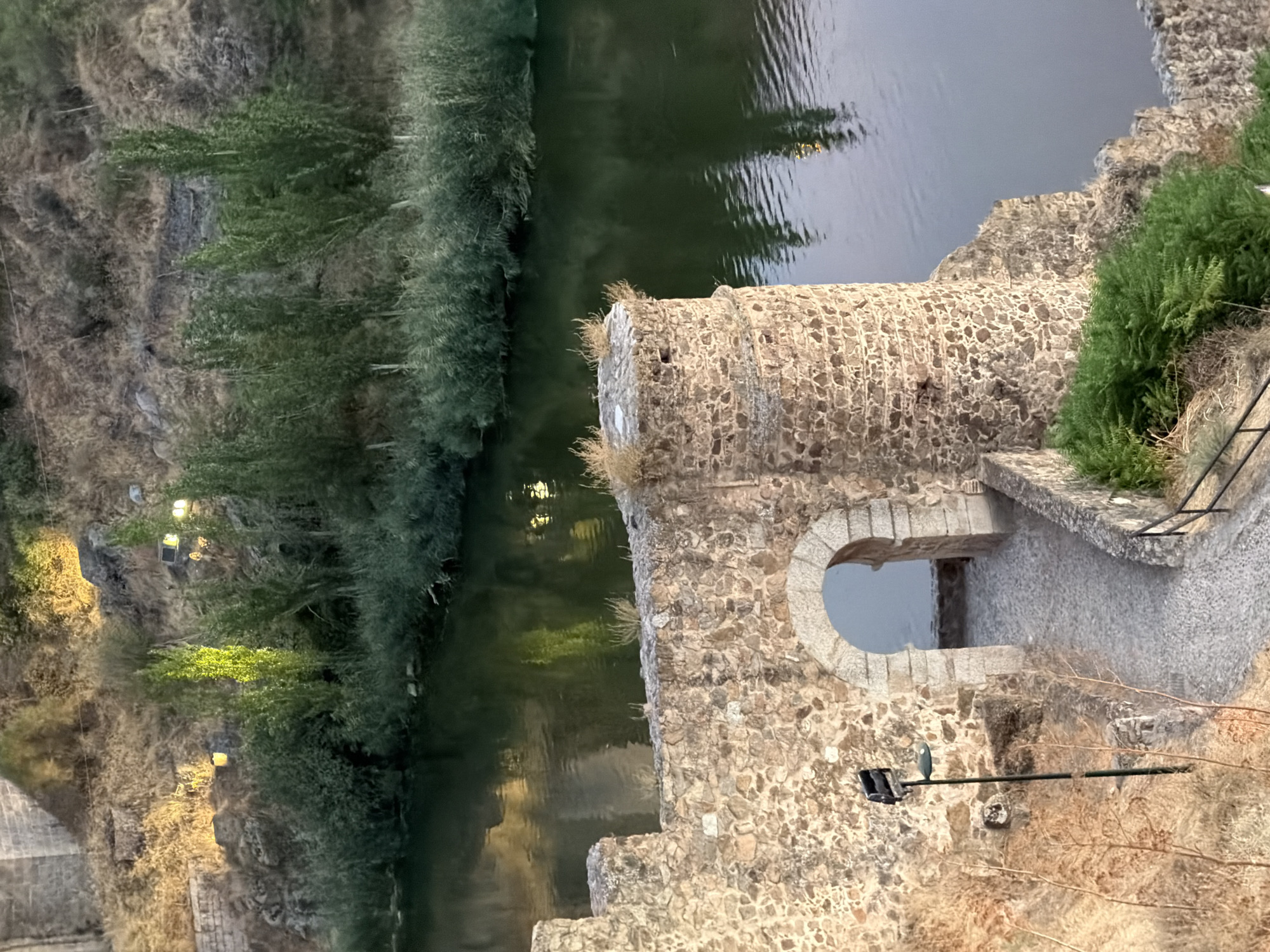 An exterior, high-angle medium shot captures a river flowing past an ancient stone fortress in Toledo, Spain. The fortress wall, constructed of rough, uneven stones, features an arched entryway leading to a cobblestone path. A cylindrical turret rises on the right side of the wall. Green vegetation flanks the river and the fortress. The river's surface reflects the surrounding trees and some artificial lights, suggesting it is late evening or early morning. The background shows a dry, rocky hillside with sparse vegetation, illuminated by several strategically placed spotlights, highlighting the historical architecture of the city.
