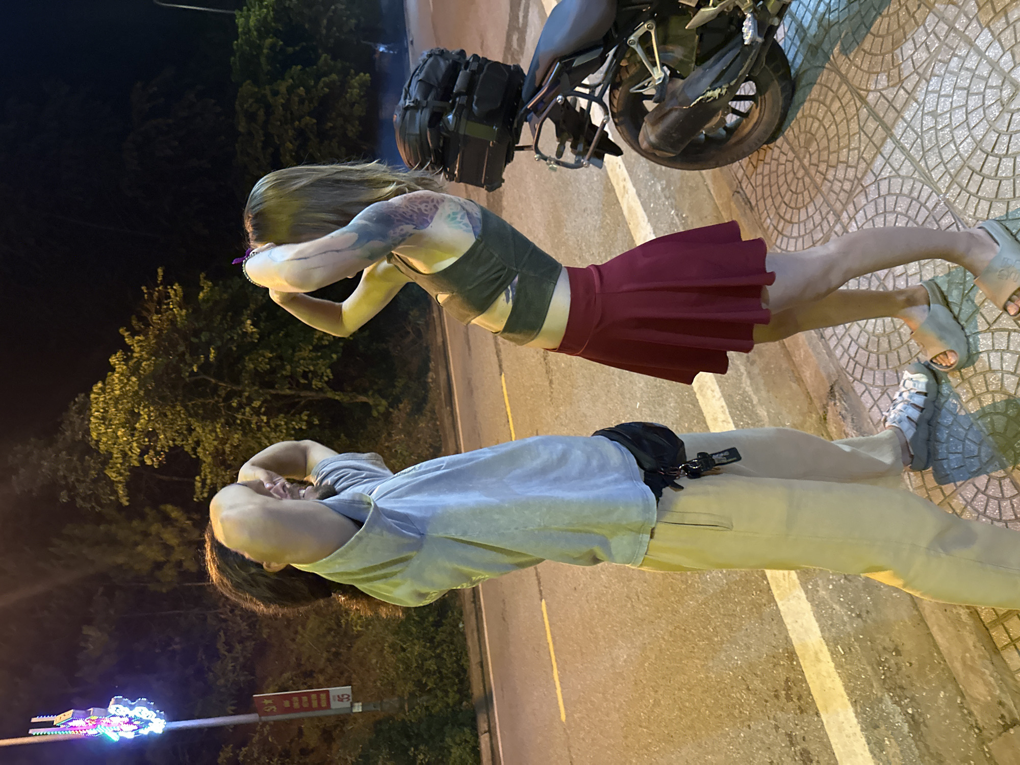 Two individuals are captured at night, standing next to a paved road in what appears to be Mù Cang Chải, Vietnam. The scene is illuminated by artificial light from the left, casting shadows.
The person in the foreground has long, light brown hair and is wearing a dark green or grey crop top and a maroon pleated mini skirt. Visible tattoos adorn their arms and shoulders. They are wearing light-colored slide sandals. Next to them, a black motorcycle with luggage strapped to its rear is partially visible.
The second person, slightly behind, has dark hair and is dressed in a light blue or grey short-sleeved top, light beige or khaki pants, and light-colored chunky sandals. A black fanny pack is worn around their waist.
Both individuals are in a similar pose, with their arms raised and bent, hands near their heads, suggesting they are stretching, posing for a photo, or engaged in a casual activity.
The setting includes a tiled sidewalk to the right, a paved road with a yellow line to the left, and lush, dark foliage in the background, indicating a natural or semi-rural environment. On the far left, a signpost features a colorful, illuminated decorative element and some unreadable red text on a white background, with the number '5' being discernible. The overall atmosphere is dark, typical of nighttime, with specific areas brightly lit by streetlights.