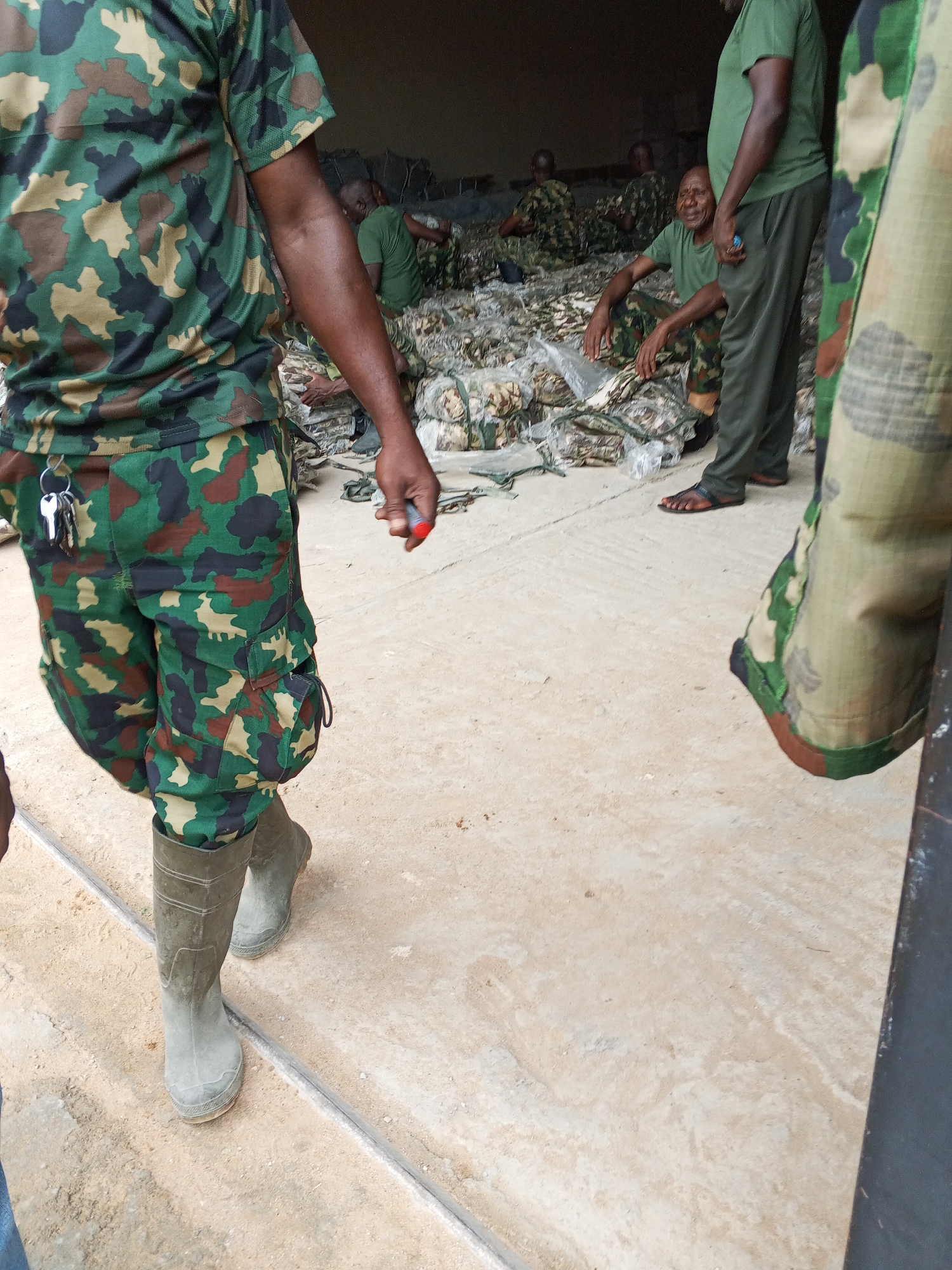 In Pasere, Nigeria, a group of soldiers are seen gathered in a room with a concrete floor. Some soldiers are seated, while others stand. The soldiers wear camouflage uniforms and rubber boots. Many camouflage packages lay on the ground.