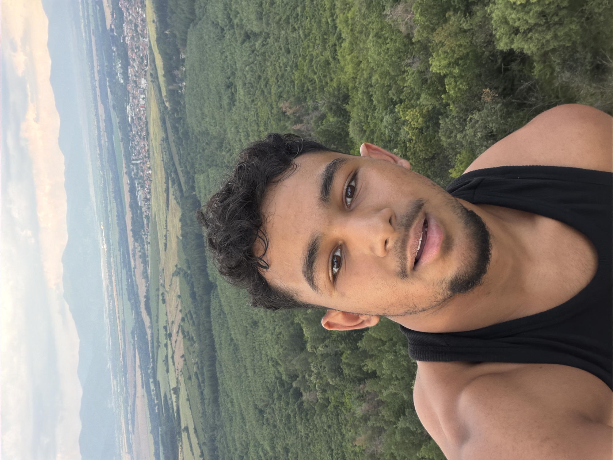 This media file is a vertical, medium shot selfie of a young man with dark, curly hair and a beard, taken outdoors. He is wearing a black tank top and is looking at the camera with a slight smile. His eyes are dark brown, and his skin is tanned.
The background reveals a sweeping landscape. In the distance, a town or village is visible with red-tiled roofs, nestled between green fields and a dense forest. Mountains can be seen in the far background, under a hazy, light-colored sky. The overall impression is of a rural or mountainous setting, likely during daylight hours with fair weather.
The man appears relaxed and happy, enjoying the view or the moment. The image seems to capture a personal moment of enjoyment and connection with nature. There is no visible text in the image. The location context provided is Turiya, Bulgaria.