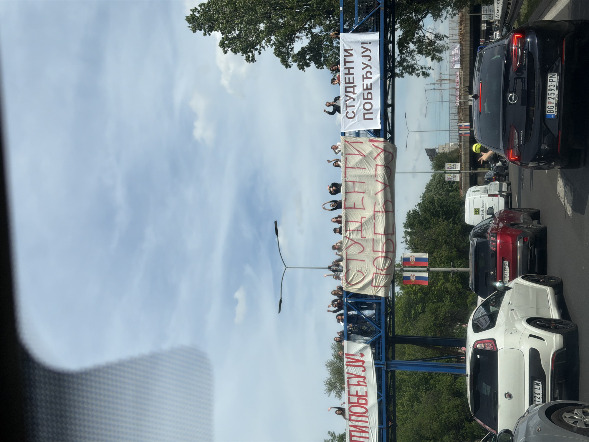 The image shows a traffic jam on a road in Belgrade, Serbia. A pedestrian bridge is visible in the background, with people holding up banners. The banners read "STUDENTS WIN!" and "THEY WIN!". The traffic jam consists of several cars, including a black Opel Insignia with license plate BG 2593-PN, a white Fiat, and a red Peugeot. A van belonging to "RanSek trade" is also visible. Serbian flags are displayed near the pedestrian bridge. The photo was taken from within a car.