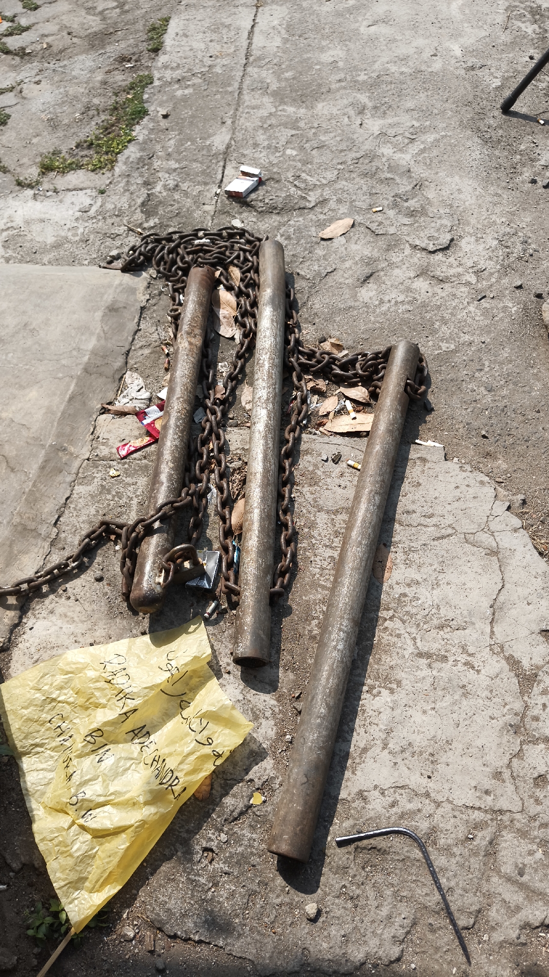 The image depicts a collection of rusty metal poles and chains lying on a cracked concrete surface, likely an outdoor area. Several discarded cigarette butts and pieces of crumpled paper, including a yellow sheet with handwritten text, are scattered around the objects. The text on the yellow paper appears to be in Arabic script. The overall scene suggests an outdoor, somewhat neglected space. The presence of broken concrete and debris indicates it might be a construction site, an abandoned area, or a general urban outdoor space. The sunlight casts distinct shadows, indicating a bright, sunny day.