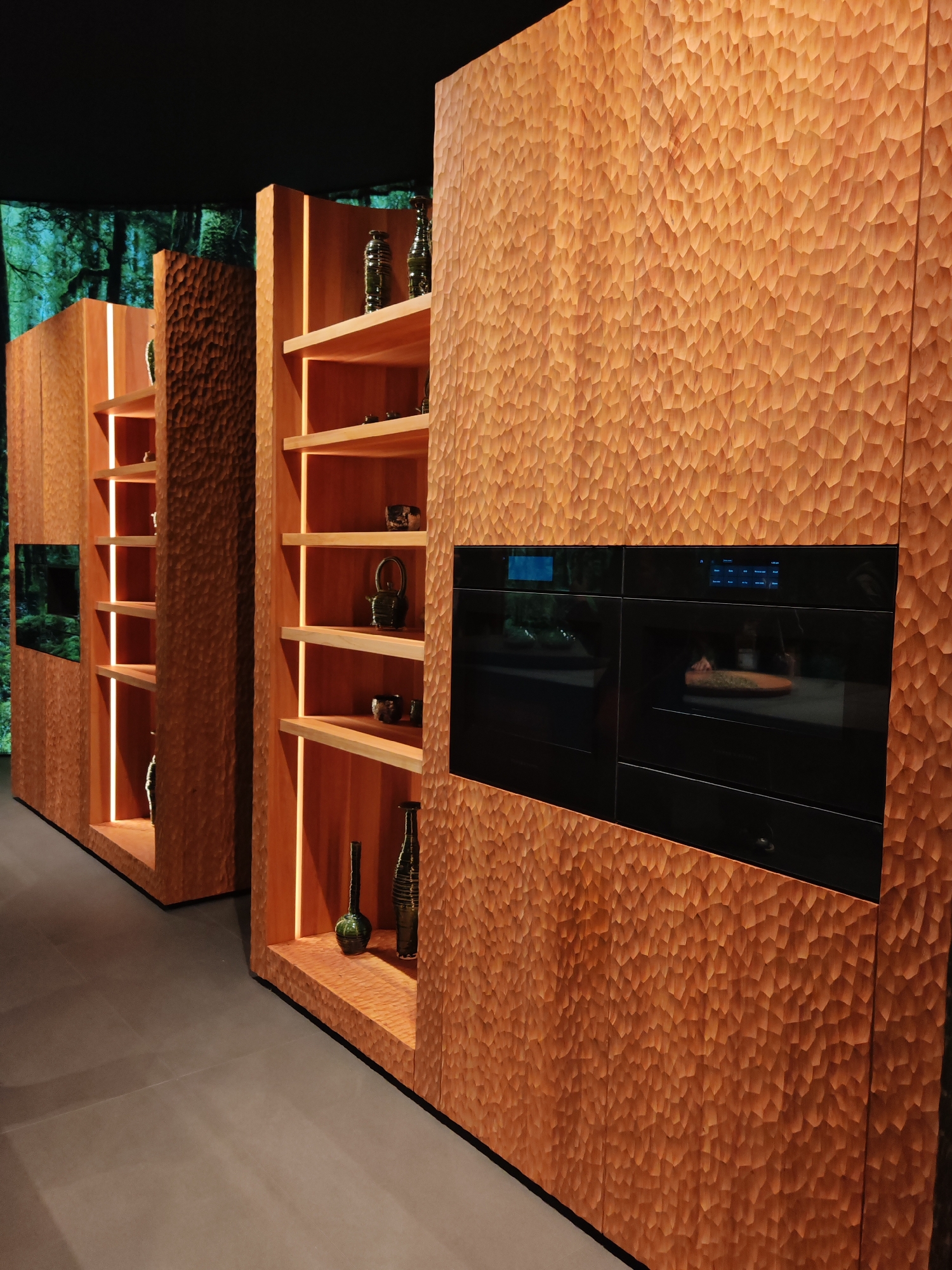 The image displays an interior space featuring extensive wooden cabinetry with a distinctive, deeply textured surface. This texture appears to be hand-chiseled or carved, creating a mosaic of small, irregular facets that reflect light unevenly, giving the wood a rugged, organic, and tactile appearance.
Integrated into these wooden structures are several open shelves, each illuminated by warm, linear LED strips embedded vertically along the sides. These shelves display a collection of decorative ceramic and glass objects, including dark-colored vases, teapots, and bowls, presented sparsely to highlight their forms.
On the right, a large section of the textured cabinetry seamlessly incorporates two modern, built-in ovens or similar kitchen appliances. These appliances have sleek, dark, reflective glass fronts; one features a blue digital display showing icons and text, while the other reveals a dish or object inside its illuminated interior.
The background features a large projection or screen displaying an image of a dense, verdant forest, adding a natural and expansive element to the indoor setting. The floor is a solid, dark grey surface, providing a neutral base that grounds the richly textured wood.
The scene suggests a high-end kitchen or a display area in a showroom, blending rustic natural materials with contemporary technology and lighting design. The overall aesthetic is one of sophisticated naturalism and calm. No people or activities are visible, presenting a static, artfully arranged space. The time of day is indeterminate due to artificial lighting. Text on the appliance displays is present but not clearly legible.