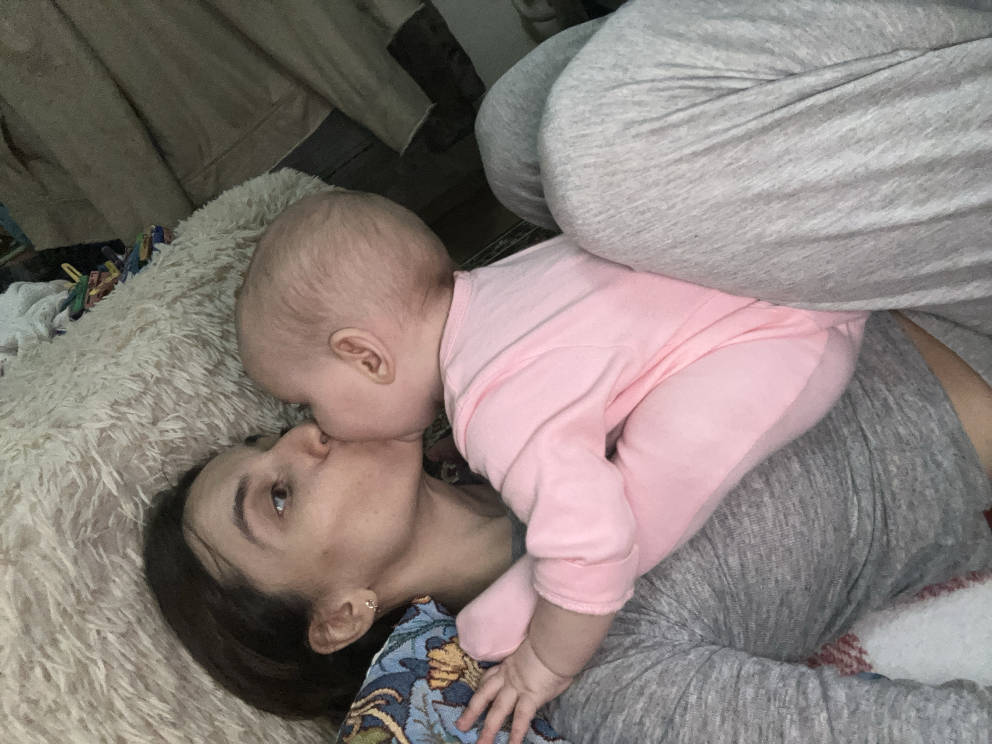 A mother and baby are lying on a bed or floor. The mother is on her back, with her head resting on a fluffy, light-colored surface. She has dark hair and is wearing a gray top with a floral-patterned fabric visible near her shoulder. The baby, dressed in a pink onesie, is positioned on top of the mother, appearing to kiss her on the face. Someone in gray pants is in the background of the shot. The location is not known but based on the provided context, it is not possible to determine if this photograph was taken in Moscow, Russia.