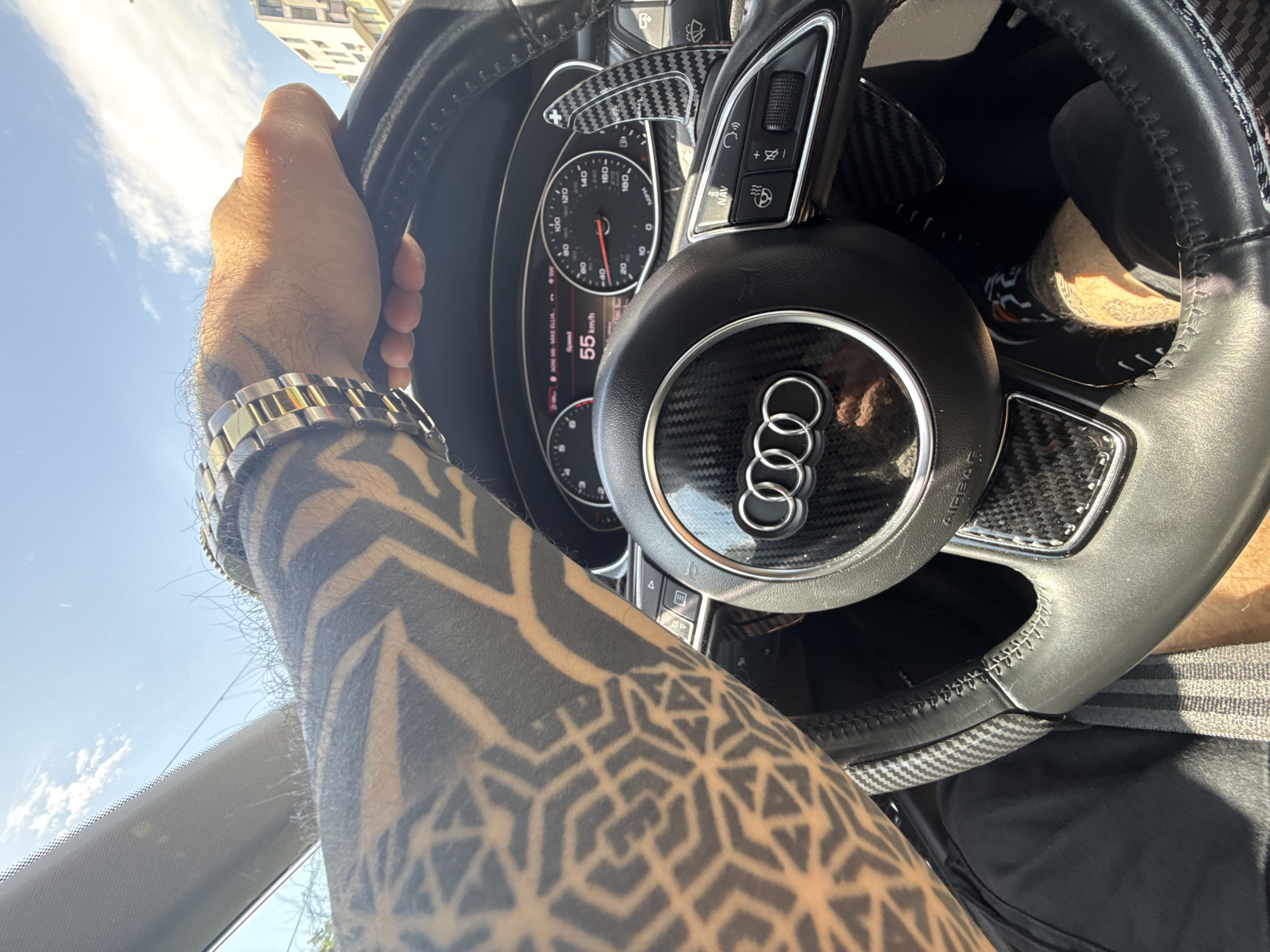 A first-person perspective image shows an individual driving a car, identified as an Audi, based on the prominent logo on the steering wheel. The driver's left arm, heavily tattooed with geometric patterns, is visible holding the steering wheel. A gold and silver wristwatch is on the driver's wrist. The car's dashboard and instrument panel, including the speedometer indicating a speed of 55 km/h, are partially visible. The exterior view through the windshield reveals a bright, sunny day with a building partially visible in the top left. The city of Sofia, Bulgaria, is not directly identifiable from visual elements in the image.