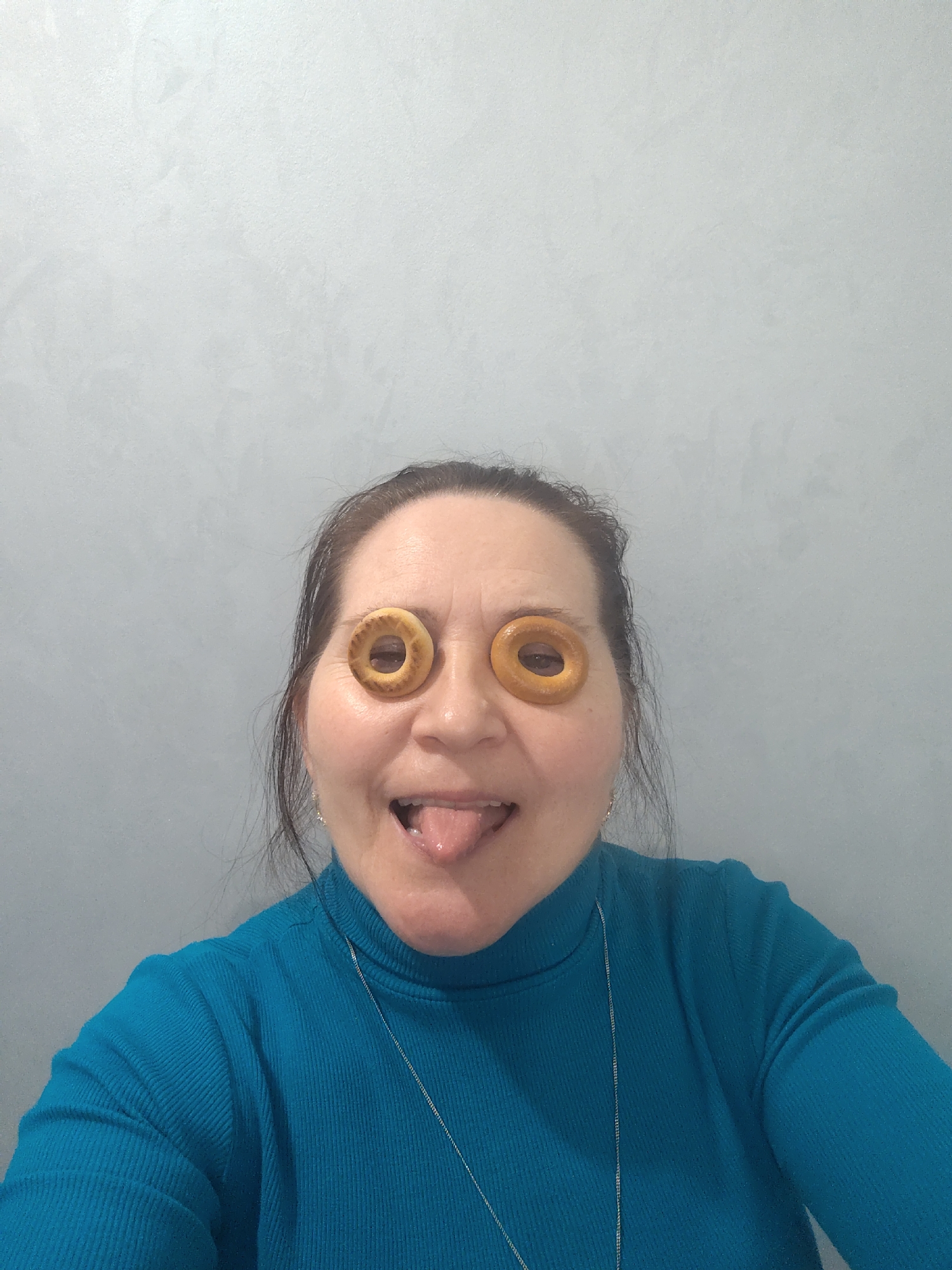 A woman is captured in a playful, close-up, indoor shot, humorously posing with two small, ring-shaped, light brown baked goods placed over her eyes. She is smiling broadly with her mouth open and tongue extended, conveying a cheerful and comical mood. She wears a ribbed, teal-colored turtleneck shirt and a thin silver chain necklace is visible around her neck. Her dark hair is pulled back from her face. The background consists of a plain, light grey wall with a subtle textured finish. No text is visible in the image.