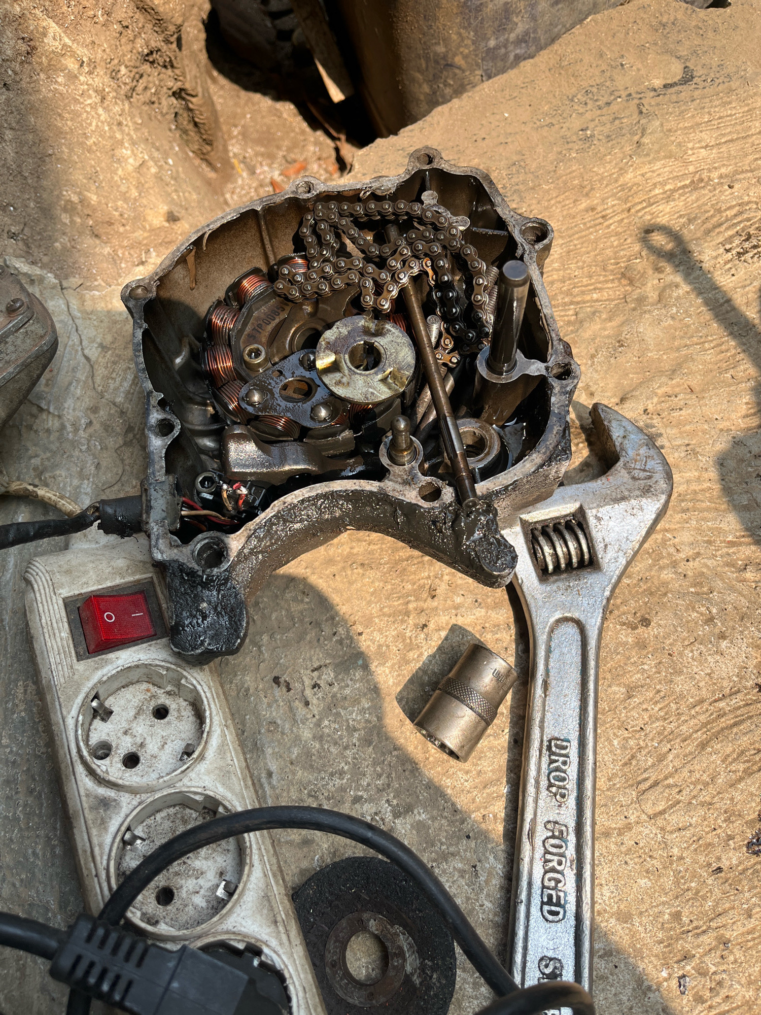 An overhead, close-up shot captures a disassembled motorcycle engine casing and various tools on a rough concrete surface. The engine casing, made of a dark metal and showing signs of wear and oil residue, is open to reveal internal components including copper wiring, a metal chain, and various bolts and gears. To the right of the engine, a large, adjustable wrench with "DROP FORGED" clearly stamped on its handle rests on the concrete. A smaller metal socket sits nearby. In the lower left, a white power strip with multiple outlets and a prominent red ON/OFF switch is visible. A black power cord snakes from the strip. The lighting suggests an outdoor environment, possibly a workshop or roadside repair area in Kebumen, Indonesia, indicated by the presence of these common repair items. The background is blurred, showing a sandy or earthy texture, further supporting an outdoor setting.