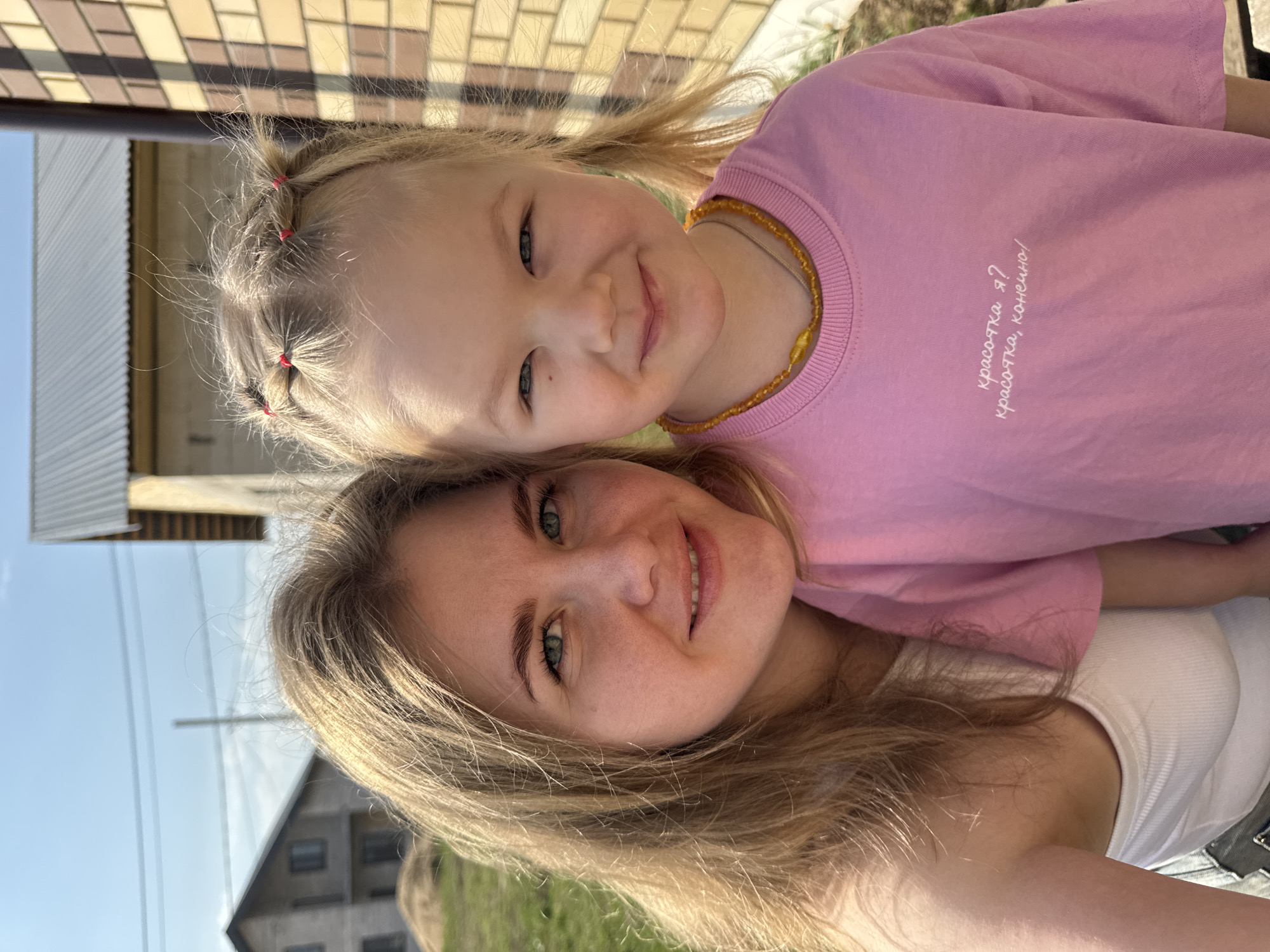A young girl with blonde hair in pigtails and a woman with long blonde hair pose for a selfie in Ферма, Russia. The girl is wearing a pink t-shirt with text in Russian, and a necklace made of amber-colored beads. The woman is wearing a white tank top. Behind them is a house with brick and corrugated metal siding and a blue sky.