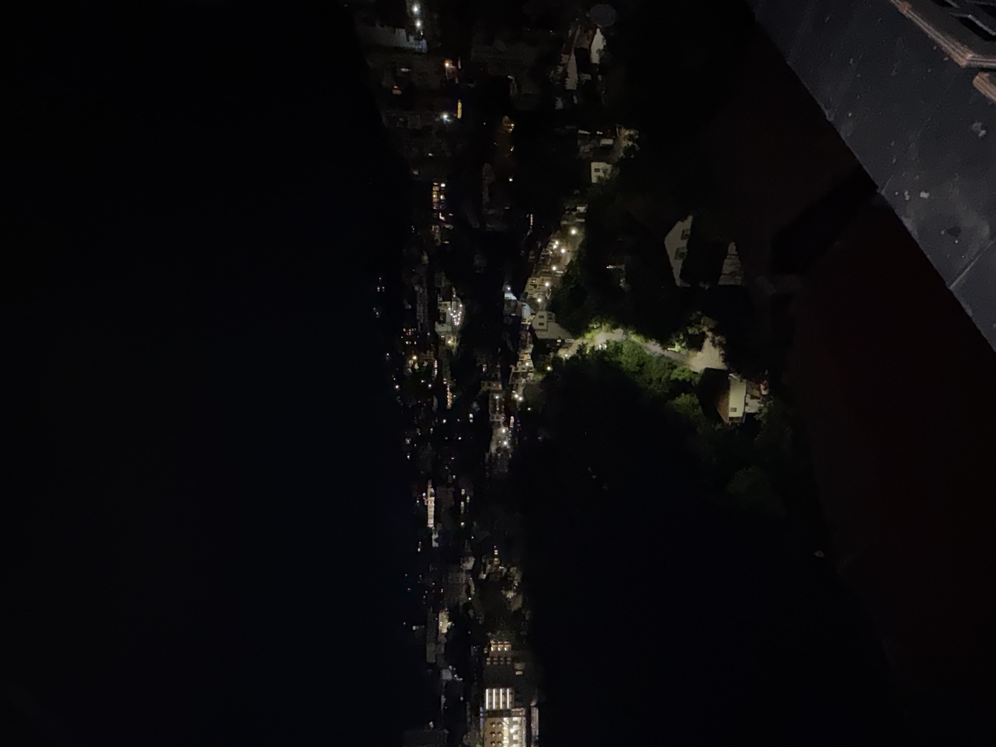 This is a night-time view of a city, likely Veliko Tarnovo, Bulgaria, given the information provided. A body of water, possibly the Yantra River, occupies the left side of the frame, appearing as a large, dark expanse. On the right side is a view into the city, with a street lit by streetlights, running away into the distance. Buildings and vegetation are visible, with some illuminated windows. The top right corner shows a dark structure, possibly a rooftop or balcony from where the photograph was taken. The scene is dimly lit, with varying degrees of darkness throughout the image.