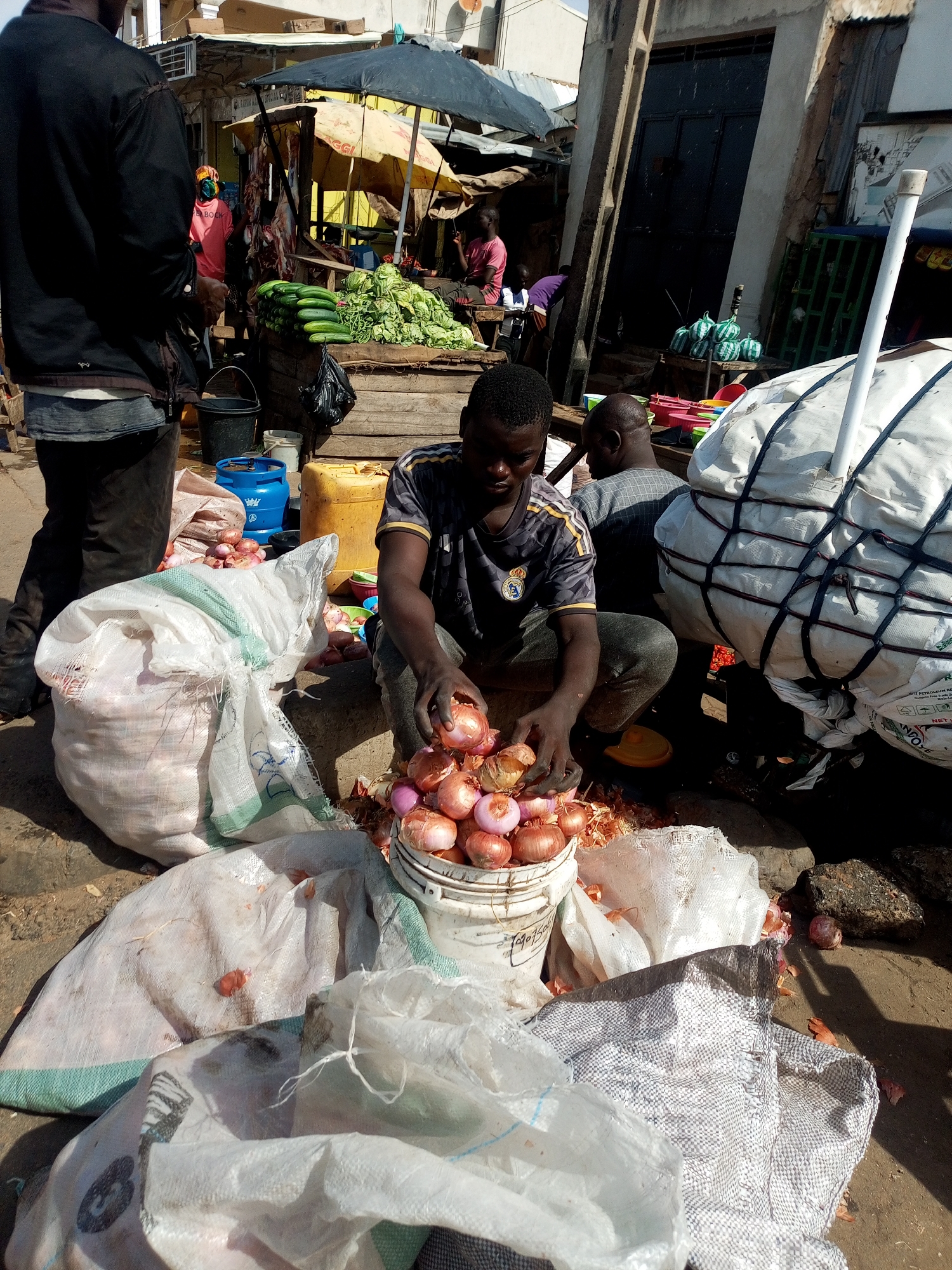 This image depicts a bustling outdoor market scene in Tempure, Nigeria, bathed in bright, direct sunlight, evident from the strong shadows cast.
In the foreground, a young Black man, wearing a dark grey football jersey with a Real Madrid crown logo, is crouched, actively sorting reddish-brown onions. He is peeling some onions, with the discarded skins scattered on the dusty ground around a white plastic bucket filled with the sorted produce. Nearby, several large white woven bags, some full of onions, are resting on the ground. To his right, a large, bulky white bag, secured with black straps, stands upright; faint text on it includes "PETROLEUM REFINERY..." and "NDOLE." Another man is partially visible to the right, also in a crouched position.
To the left, a person in a black jacket is seen from behind, walking or standing. In the mid-ground, a market stall under a yellow umbrella, possibly branded "MAGGI" (with "GGI" visible), displays piles of fresh green produce, including cucumbers and leafy greens. Below the stall, what appears to be hung meat is also visible. Another person in a bright pink shirt with "BOCKY" written on the back stands near the stall. Various market items like black buckets, a blue gas cylinder, a yellow jerry can, and colorful plastic containers are scattered on the ground. The background features simple concrete buildings and the general hustle of an active open-air market.