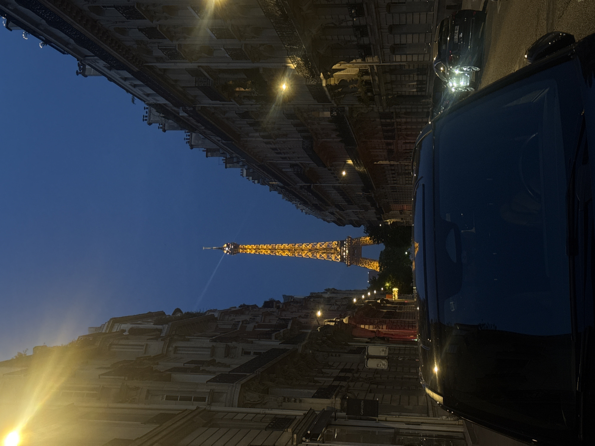 The image captures a view down a narrow street in Paris, France, at dusk or night, with the Eiffel Tower illuminated in the distance. The street is flanked by tall, classic Parisian buildings with multiple stories, windows, and balconies. Streetlights are visible, casting a warm glow on the scene. In the foreground, the hood and windshield of a black car are prominent, suggesting the photo was taken from inside or directly behind a vehicle. Another car, with its headlights on, is visible further down the street. The sky is a deep blue, indicating the time of day. The Eiffel Tower, the iconic landmark of Paris, stands tall and brightly lit, serving as the focal point of the image.