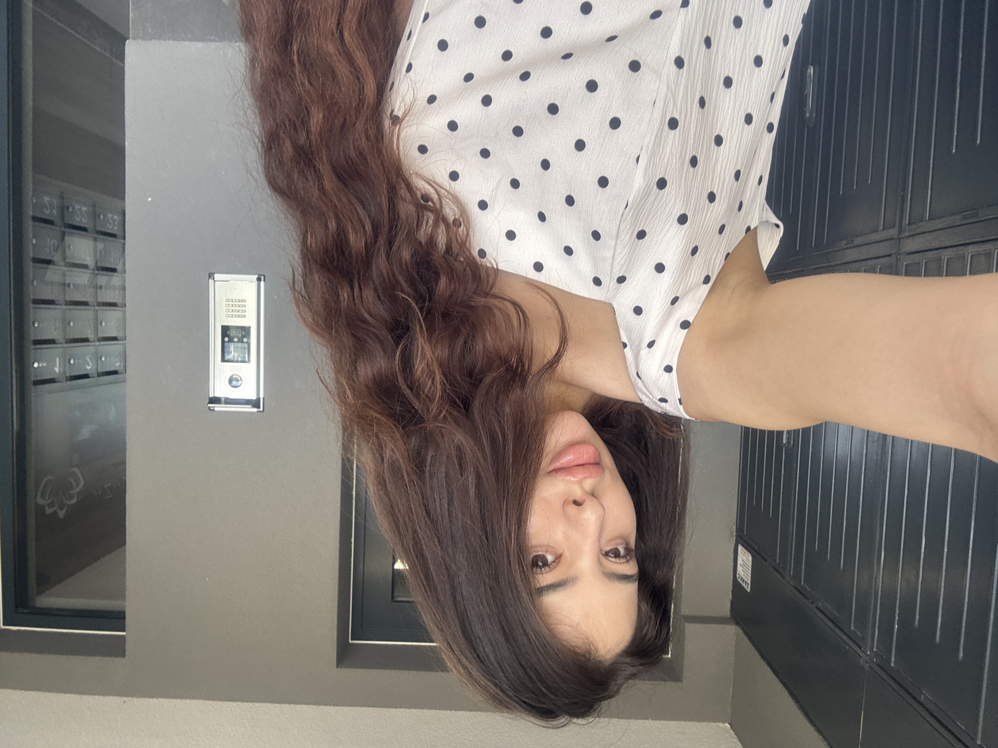A woman with long, wavy brown hair, wearing a white blouse with black polka dots, is taking a selfie. She is standing outside a building. Behind her are mailboxes, an intercom system, and a dark grey metal door. The location is likely an apartment building. Due to a lack of information in the image, the exact location within Plovdiv, Bulgaria cannot be determined.