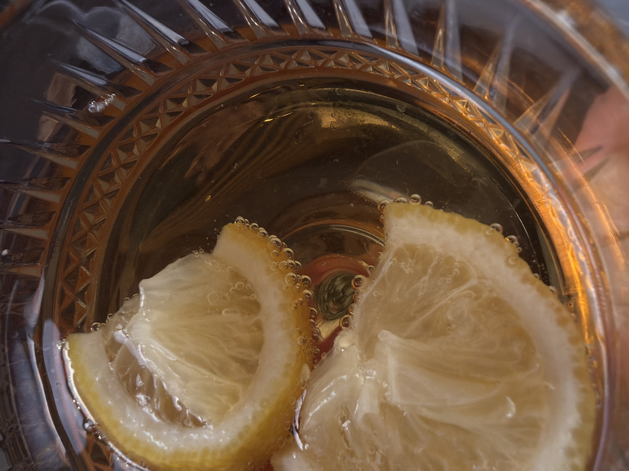 The image depicts a close-up, overhead shot of a clear, ornate glass filled with a light-colored liquid and two slices of lemon. Small bubbles cling to the lemons and the glass. The glass itself has a patterned design around the rim. No city or location is discernible from the image.