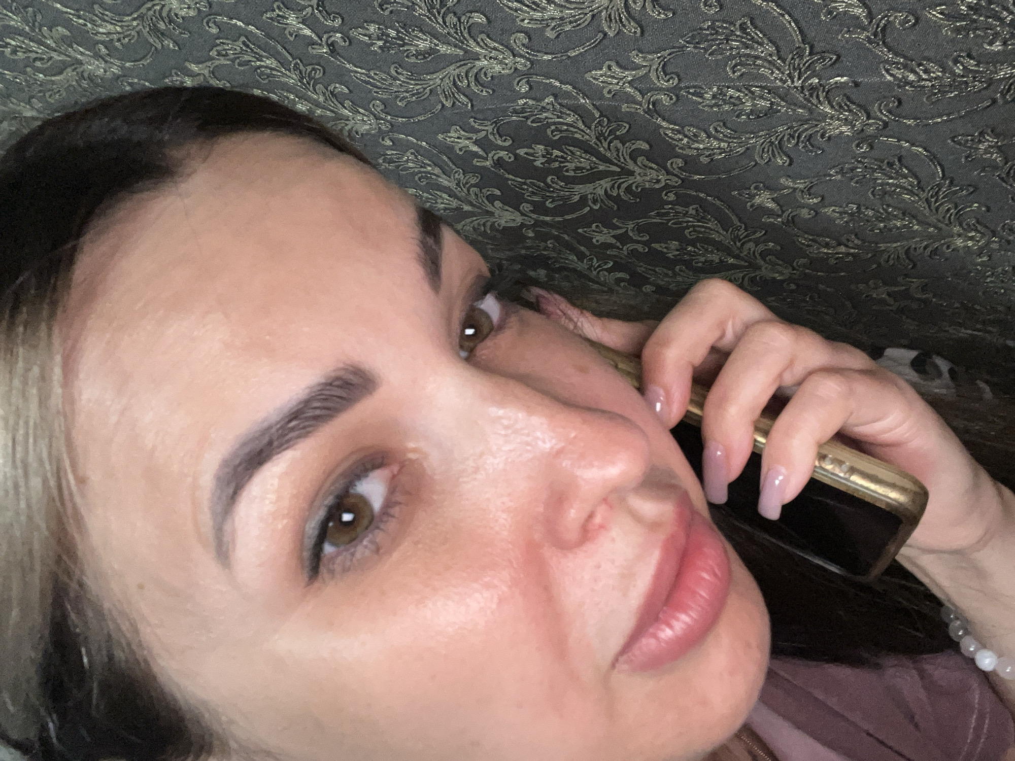 A close-up, eye-level, indoor shot shows the face of a woman as she holds a gold-colored cell phone to her right ear. The woman is facing the camera, with her head tilted to her left and resting against a dark gray, patterned fabric surface. Her left eyebrow is arched, and her eyes are open and looking at the camera. She has fair skin with some redness on her cheeks, full lips, and dark hair pulled back from her face. Her fingernails are long, square-shaped, and painted with a sheer pink polish. The phone's screen is dark, and the top edge of the device is visible. The background is a textured, damask-patterned fabric in shades of gray and black. The lighting is soft and appears to be coming from the front, illuminating the woman's face and the phone.