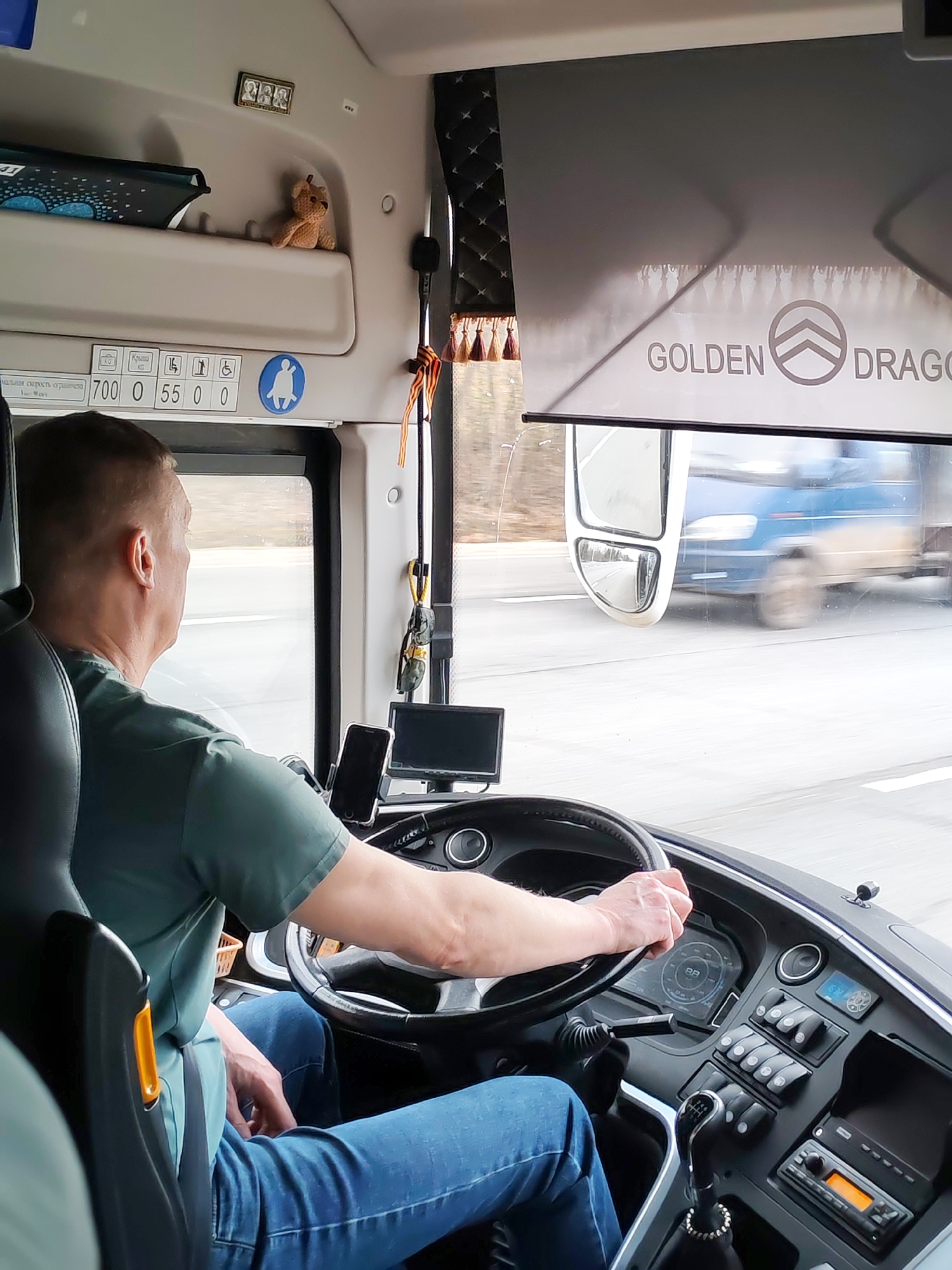 The image captures the interior of a bus cockpit from a passenger's perspective, looking forward. A male bus driver, seen from behind, is wearing a green t-shirt and blue jeans, with his hands on the steering wheel. The bus appears to be in motion on a multi-lane highway, with the outside view showing a blurred passing blue vehicle (likely a truck or another bus) through the windshield and side mirror.
The bus cabin is well-equipped with a detailed dashboard featuring a manual gear stick, various control buttons, a radio/cassette player, and digital displays. A mobile phone is mounted in a holder, and a small monitor screen is visible to the driver's right. Overhead, a beige compartment holds a small light brown teddy bear and a miniature framed picture containing three portraits. A dark blue tablet is also stored in this compartment.
A grey sunshade is partially pulled down, displaying the text "GOLDEN DRAGON" and a stylized logo. Near the side window, a decorative curtain with tassels and a St. George's ribbon are visible. Above the driver, on an interior panel, there are numbers and icons: "700 0 55 0 0", "КРЫМ 0" (Krym 0), and several pictograms indicating standing capacity, elderly/disabled access, and a blue circular icon depicting a person with a child.
The time of day appears to be daytime, with a clear, though possibly overcast, sky. The driver appears focused on the road, suggesting a routine journey. The presence of the St. George's ribbon aligns with the given location context of Krasnyi Bor, Russia, and is a common cultural accessory in that region.