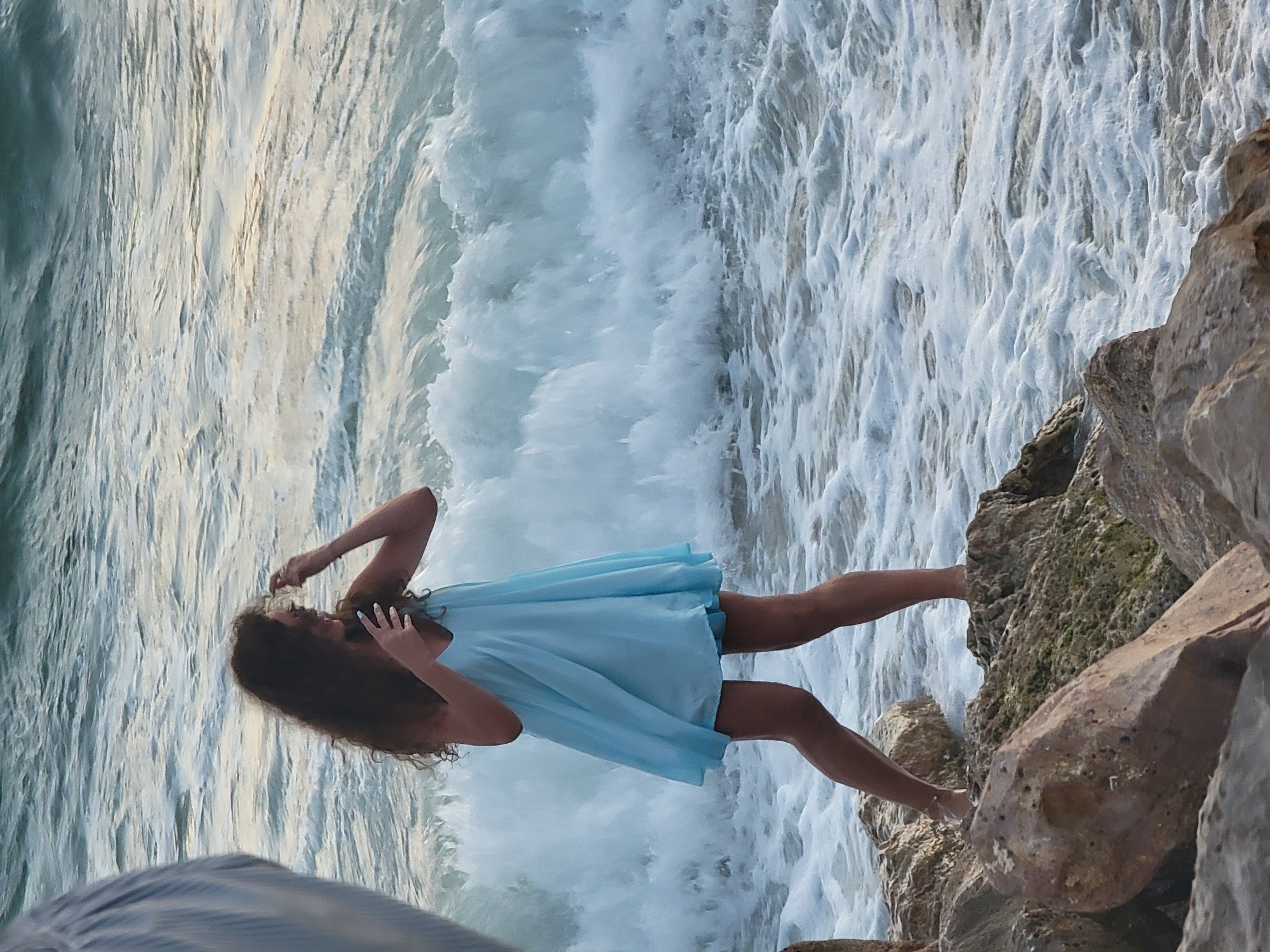 A woman with long, curly, dark hair is standing on rocks near the shore, wearing a light blue, flowing dress. She is facing the ocean, with waves crashing behind her. The water is a mix of blue and white foam, and the sky appears overcast. The scene is captured from a slightly elevated perspective, with the foreground dominated by large, textured rocks. The location is the rocky shoreline of Quarteira, Portugal.