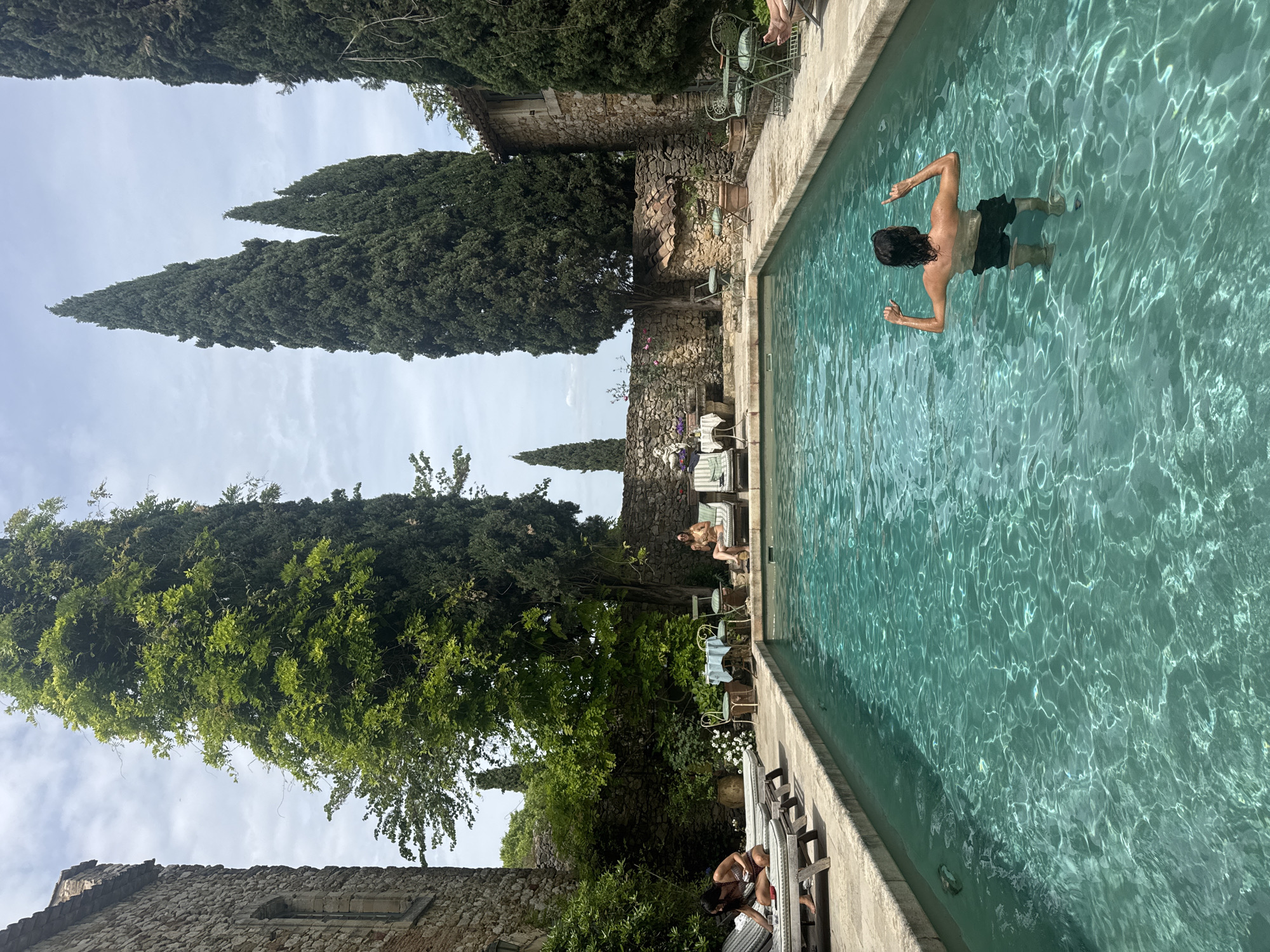 The image depicts a rectangular swimming pool in Saint-Victor-la-Coste, France. A man with long dark hair and black swim shorts is in the pool with his arms raised. Two people are sitting on lounge chairs along the pool's edge. A stone wall borders the area with tall, narrow trees interspersed along the perimeter. Other chairs and tables are present on the poolside.