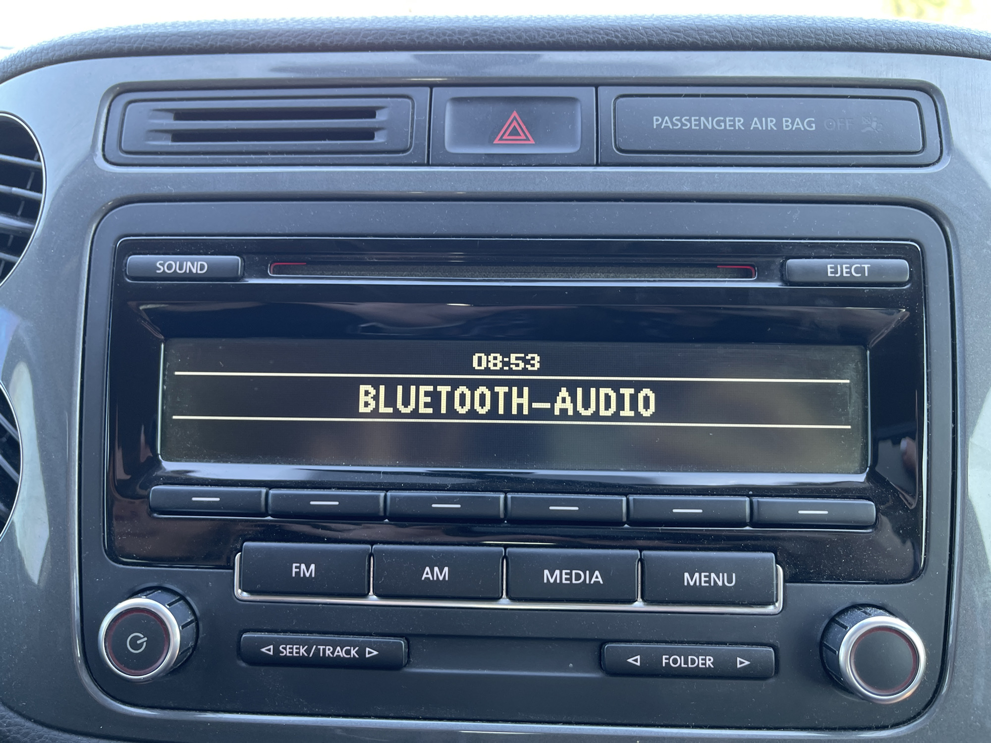 The image shows a close-up view of a car's central console, specifically the infotainment system and surrounding controls. The screen displays "08:53" and "BLUETOOTH-AUDIO" in pixelated text. Below the screen are several buttons labeled "SOUND", "EJECT", "FM", "AM", "MEDIA", and "MENU", along with directional arrows for seeking tracks or folders. A circular power button with a 'G' symbol is visible on the left, and a similar dial is on the right. Above the screen, there are air vents, a hazard light button with a red triangle symbol, and a "PASSENGER AIR BAG OFF" indicator. The console is dark grey and appears slightly dusty. The scene is devoid of people or any elements that would indicate its location in Chervonyi Lan, Ukraine.