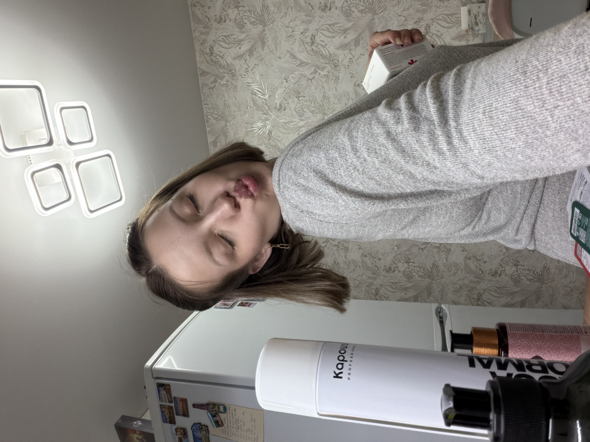 A photograph taken indoors in Reshetnikovo, Russia, shows a young woman with shoulder-length brunette hair and fair skin. She is wearing a light gray sweater and is positioned so that her face is angled towards the camera. Her eyes are closed, and her lips are pursed in a kiss. She has simple paper clip earrings.
Behind the woman is a light-colored wall with a floral wallpaper pattern. To her left, a modern square-shaped ceiling light fixture is visible. A white refrigerator with various magnets and papers on it is in the foreground. On top of the refrigerator, there are a few hair products, including a tall white can of Kapous Professional hair spray. The woman is holding a white box with red text and logos on it in her right hand. The scene appears to be a casual, personal photograph taken indoors.