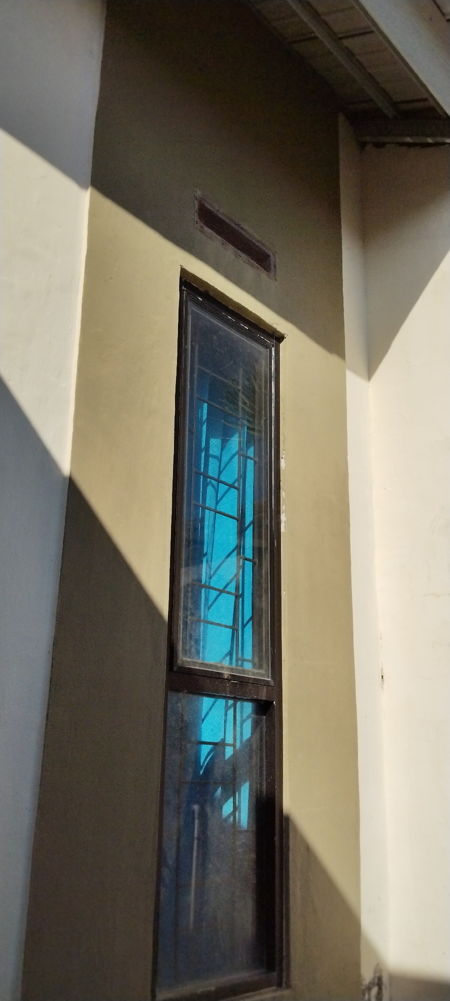 This is an outdoor, eye-level, medium shot of a narrow, vertically oriented window set into a light tan-colored stucco wall. The window has a dark brown frame and is divided into two panes by a horizontal mullion. The upper pane is filled with a bright blue glass and has a black metal grate in front of it, creating a diamond pattern. The lower pane is partially obscured by reflection and appears to be dirty. Above the window, a rectangular louvered vent is visible, also set into the wall. The wall itself is partially illuminated by strong sunlight, creating harsh shadows. To the left, a white wall is visible, also casting a shadow across the tan wall. To the upper right, the edge of a roof structure is visible, with metal joists. The scene appears to be part of a building's exterior in Duman, Indonesia. There are no people visible in the image, and no discernible actions or interactions are taking place. The overall impression is one of a simple architectural detail illuminated by natural light.