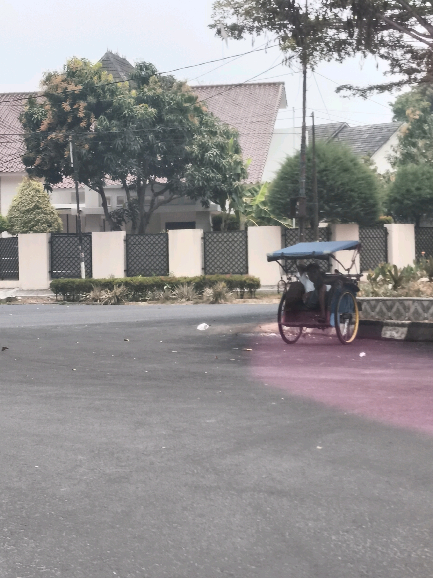 An overcast day in Palembang, Indonesia is captured in this image, showing a residential area with a prominent street in the foreground. A dark-colored, three-wheeled vehicle, commonly known as a becak, is parked on the right side of the road. The becak features a blue canopy, and a person is seated within it. The street is paved and appears to be in decent condition, with some debris scattered on its surface.
Behind the street, a beige wall with a patterned dark fence runs horizontally across the mid-ground, enclosing the properties. Beyond the fence, modern houses with tiled roofs are visible, suggesting a well-established neighborhood. Lush green trees and shrubs are interspersed throughout the scene, adding a touch of nature to the urban landscape. The sky is a pale gray, indicating cloudy weather. A patch of bright pink light is visible on the road, possibly from a reflection or artificial lighting.