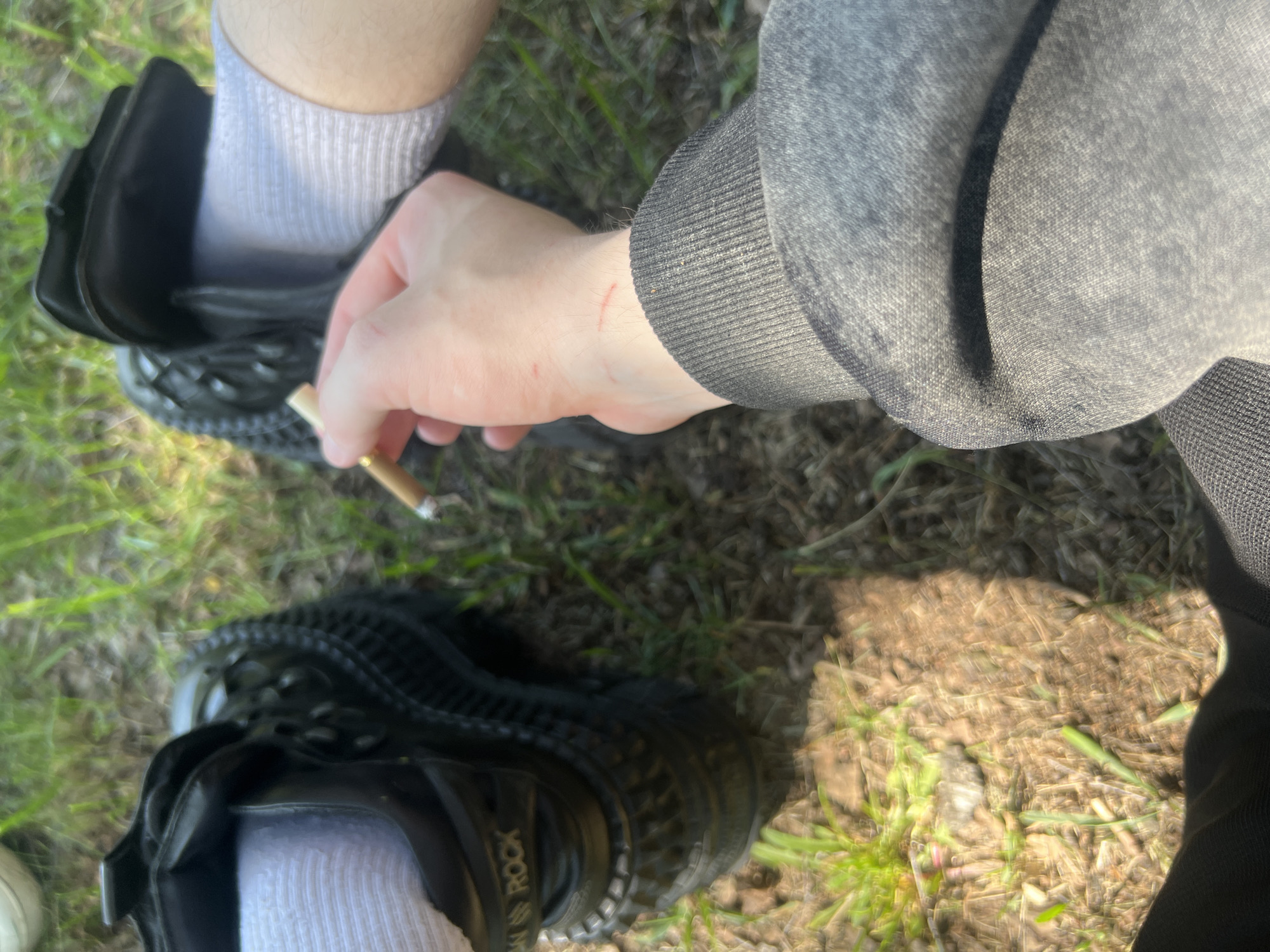 The image provides a top-down view of a person outdoors, likely in Saint Petersburg, Russia. The primary subjects are the person's lower legs, feet, and one arm and hand.
The individual is wearing white ribbed socks and black, chunky-soled shoes. The brand name "NEW ROCK" is visible on the side of one of the shoes. One foot appears fully inside a shoe, while the other is either partially inserted or being put on/taken off. The person's arm is clothed in a grey, long-sleeved top or hoodie, which features a ribbed cuff and textured fabric. The visible hand holds a lit cigarette between its fingers, suggesting the activity of smoking or preparing to smoke. Several small, reddish scratch marks are noticeable on the back of the hand and wrist area.
The setting is an outdoor environment with natural ground cover, consisting of dry grass, patches of green vegetation, and dirt. The strong highlights and shadows indicate it is daytime with direct sunlight. No specific weather conditions beyond sunniness can be inferred. The visible text is "NEW ROCK" on the side of the shoe. The scene conveys a casual, perhaps contemplative, moment outdoors.
