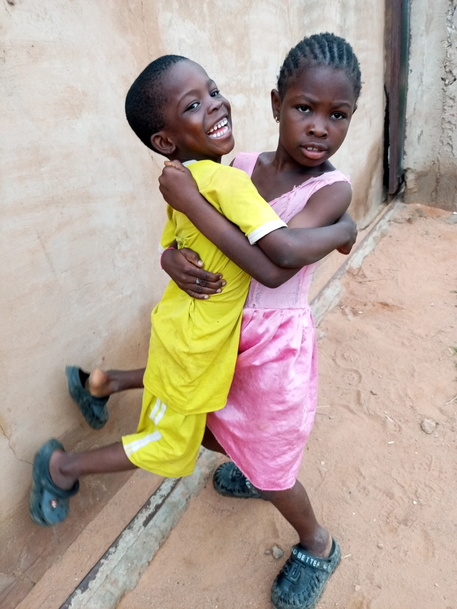 Two young Black children, one wearing a yellow t-shirt and shorts, and the other a pink dress, embrace outdoors. The child in yellow has their arms wrapped around the child in pink. They are both wearing dark clogs. The background shows a textured wall and sandy ground, consistent with the environment in Tempure, Nigeria.
