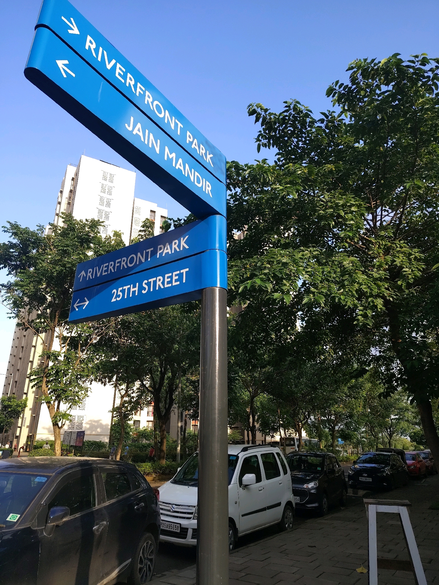 A bright blue directional signpost stands in the foreground against a clear blue sky. The sign has three levels. The top level points to the right and reads "RIVERFRONT PARK". Below that, it points to the left and reads "JAIN MANDIR". The lowest level points to the right and reads "RIVERFRONT PARK", and below that, it points to the left and reads "25TH STREET". Behind the signpost, several cars are parked along a paved sidewalk. A tall, white building with multiple windows is visible on the left, and lush green trees line the sidewalk on the right and in the background. Some pedestrians are seen walking near the building.