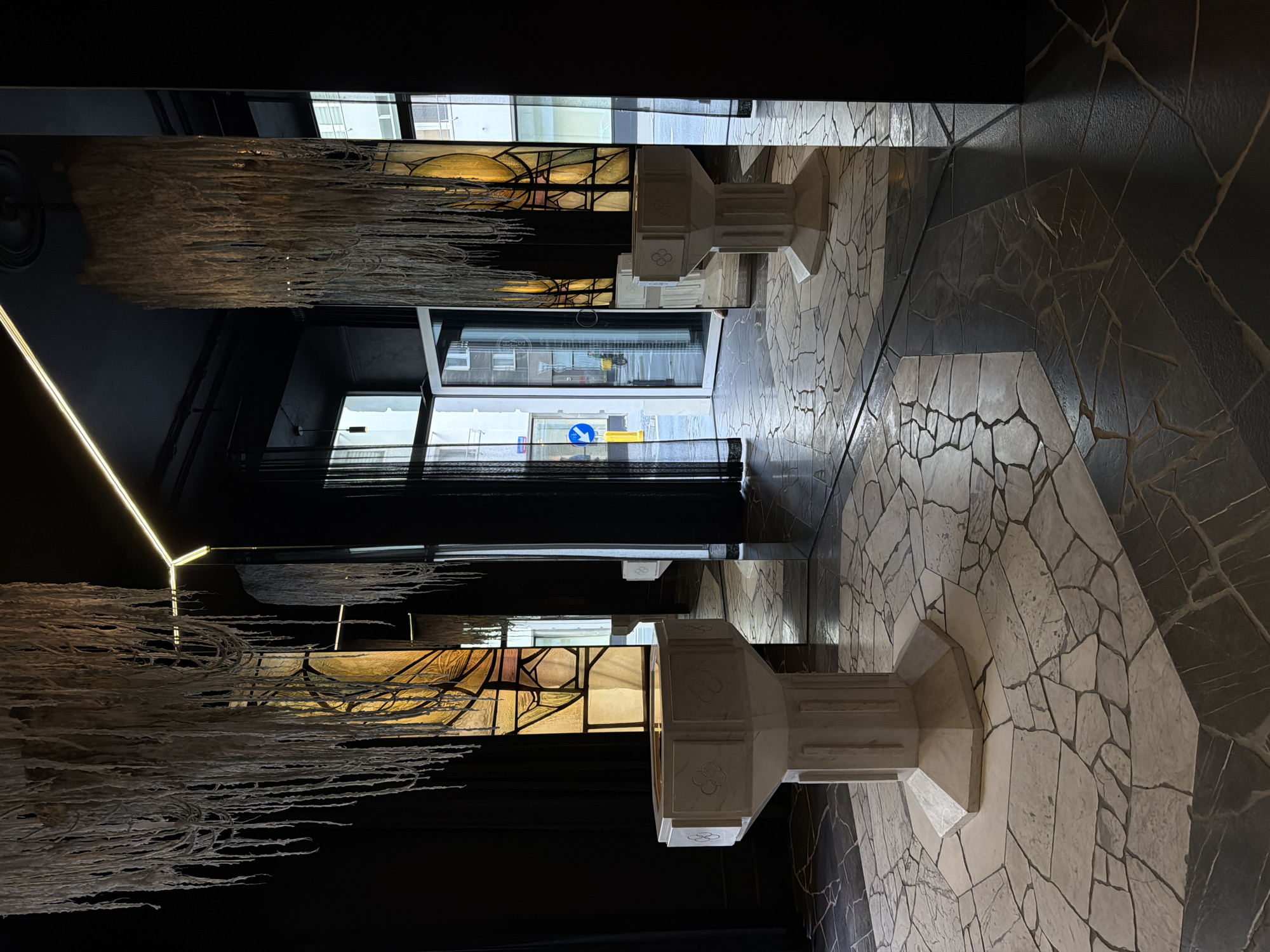 The image depicts the interior of a sophisticated, high-ceilinged space, likely a gallery, chapel, or design-conscious commercial establishment, in the city of Unknown, Unknown. The dominant color scheme is a stark contrast of dark charcoal or black walls and ceiling against lighter elements.
In the foreground, two white, octagonal pedestals, possibly baptismal fonts or display plinths, are symmetrically positioned. The prominent pedestal on the left features a fluted cylindrical base, an octagonal top with relief carvings of a clover-like motif on each side, and a recessed basin. The second pedestal is further back on the right, partially obscured and reflected in a large mirror. Both pedestals are made of a light-colored, possibly marble or stone-like material.
Above each pedestal hangs a large, cylindrical chandelier or light fixture, composed of numerous long, thin, white, fibrous strands resembling dried plant material, raw wool, or perhaps cascades of melted candle wax. These fixtures create a distinctive, organic texture against the dark ceiling.
Behind the pedestals, two tall, rectangular panels of abstract stained glass are visible, featuring patterns in shades of amber, yellow, and brown with dark leading. These panels flank an open double glass door entrance in the background, which reveals a bright, sunlit city street scene. Outside, light-colored buildings with multiple windows are visible, along with a blue circular sign displaying a white arrow pointing left, mounted on a yellow pole, and a distant pedestrian or cyclist.
The floor is a mosaic of irregularly shaped, flat stone pieces, primarily light grey and white in the central area, transitioning to darker grey and black on the sides and towards the back. The dark grout lines define the intricate, broken-tile pattern. A bright, linear LED light strip illuminates the ceiling, angled across the dark surface. Large reflective mirror panels on the walls create an illusion of extended space and provide reflections of the interior elements, including the chandeliers and pedestals. Black, sheer curtains are drawn back to frame the entrance. No people are present in the scene.