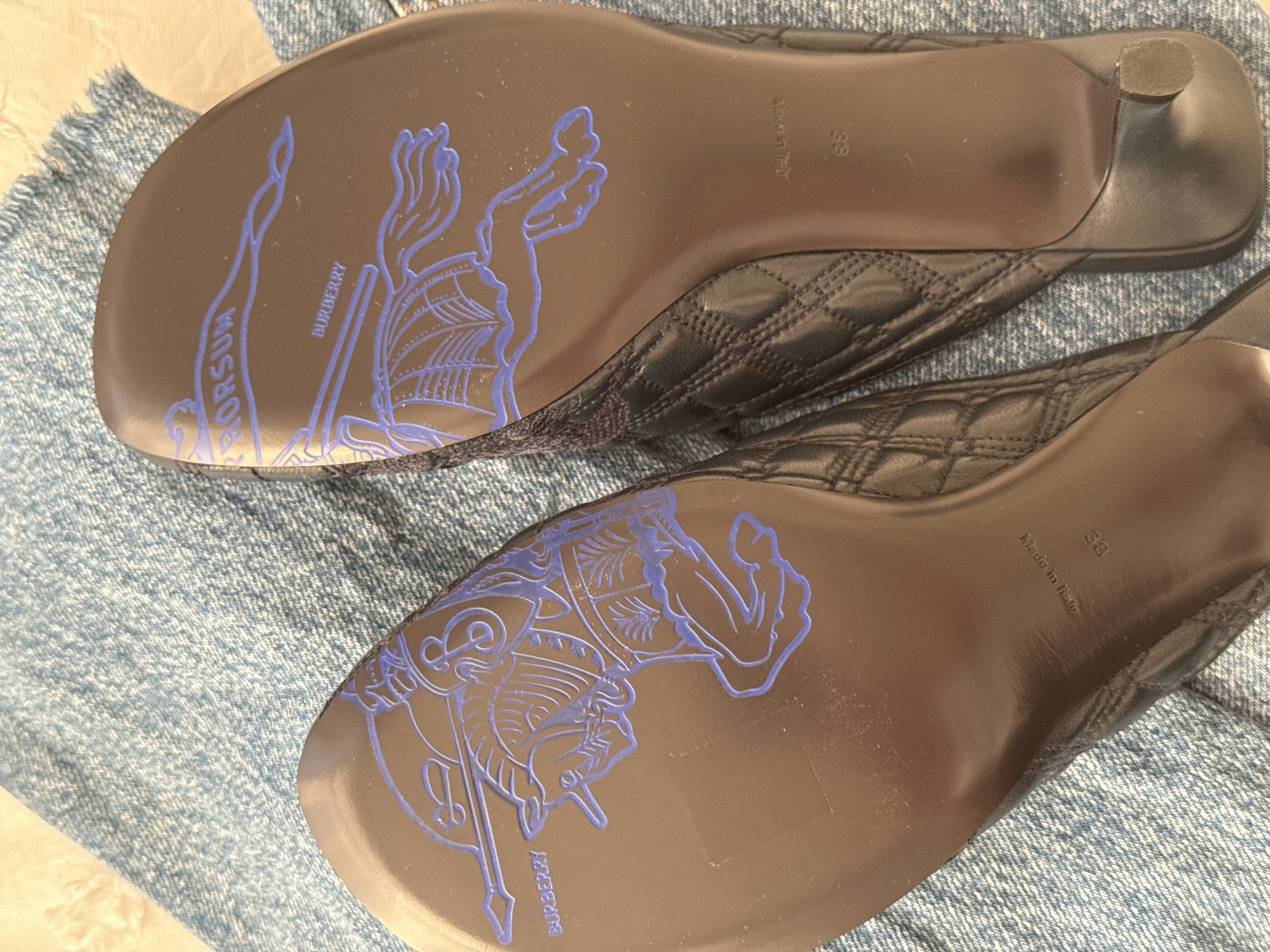 The image shows a pair of black leather mules placed on a light blue denim surface. The soles of the shoes are visible, revealing the "Burberry" logo and a depiction of a knight on horseback in blue ink. The shoes are labeled "Made in Italy" and display the size "38".