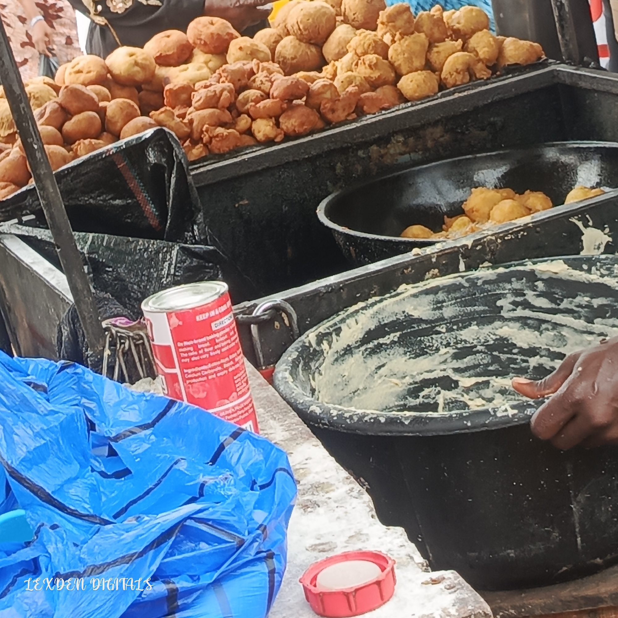 The image depicts a street food stall in Egan, Nigeria, likely in the daytime. The main subjects are piles of freshly fried dough balls, commonly known as puff-puff or bofrot. These golden-brown fritters are arranged in metal containers, overflowing with the sweet treat.
In the foreground, a large black plastic tub is filled with a pale, batter-like substance, with a dark-skinned hand reaching into it, suggesting that more dough is being prepared or fried. Beside the tub, a bright blue plastic bag is crumpled, obscuring the lower part of the stall. A red metal can with a white lid is visible next to the bag. The can has text printed on it in black and red, part of which is legible. It appears to be a food product, with instructions and ingredients listed.
Behind the food containers, parts of people are visible. Their attire suggests a casual setting, possibly a market or a busy street. The overall scene is one of food preparation and sale in a vibrant, outdoor environment. The lighting suggests it is daytime, and there are no clear indicators of specific weather conditions. The presence of the food, the preparation tools, and the people engaged in commerce create a sense of daily life and economic activity in Nigeria.