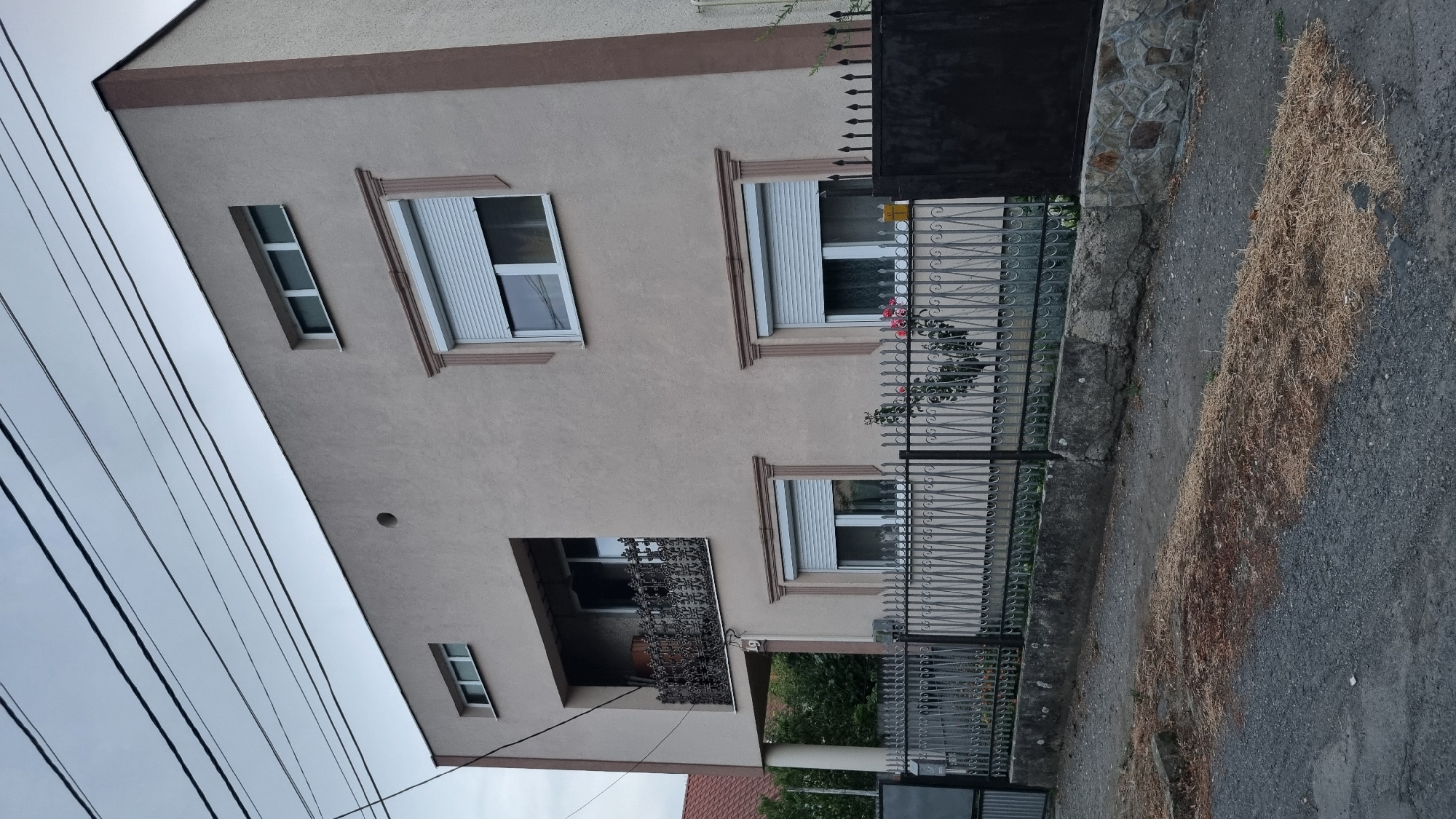 A beige, three-story building with a brown trim is viewed from a low angle on a cloudy day. The building has several windows with white shutters. A balcony with ornate iron railings is on the second floor. A black metal gate and a decorative metal fence are in front of the building. The ground in front of the fence is covered with a layer of dry grass. The building is located on a street in Berehove, Ukraine.