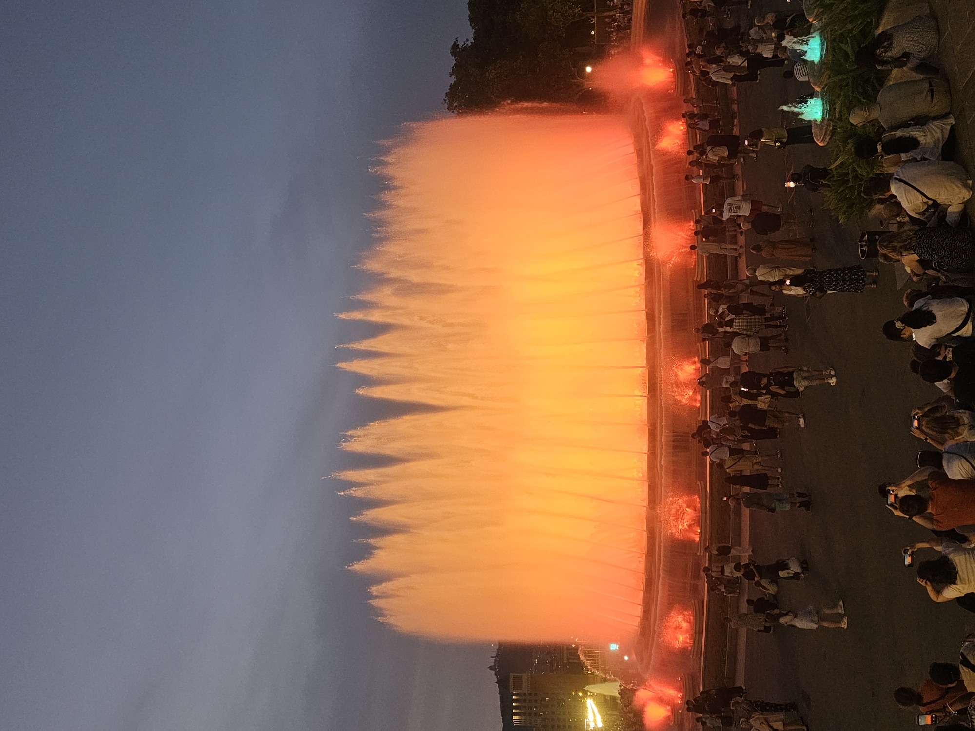 The image depicts the Magic Fountain of Montjuïc in Barcelona, Spain, during the evening. A large, illuminated fountain dominates the scene, with water jets creating a curtain of orange and yellow light against the darkening sky. The fountain is surrounded by a tiered structure with red and orange lights at its base, and smaller, green illuminated fountains are visible to the right. A crowd of people are gathered in the foreground and middle ground, observing the fountain. Many are standing, while some are sitting or kneeling. Several individuals are holding up their mobile phones, presumably to take pictures or videos. The environment appears to be an open public space with paved ground and some trees and buildings visible in the background.