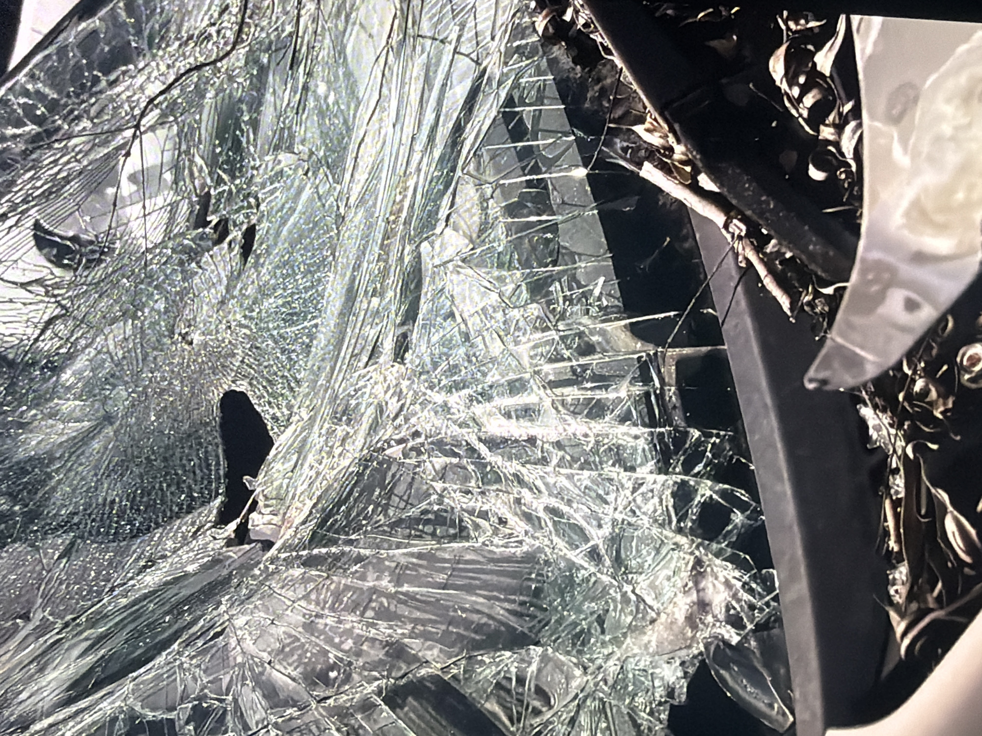 This is a close-up, high-angle shot of a severely damaged windshield. The glass is shattered into numerous fragments, creating a web-like pattern of cracks. There is a large, dark void where a significant portion of the glass is missing, exposing the interior of the vehicle. The remaining glass pieces are sharp and jagged, reflecting light and creating a distorted view of what lies beyond. Debris, possibly leaves and twigs, is visible on the dashboard below the windshield. The overall impression is one of destruction and impact. The image is undated and its location in Bukuru, Nigeria, is not visually evident from the provided shot.