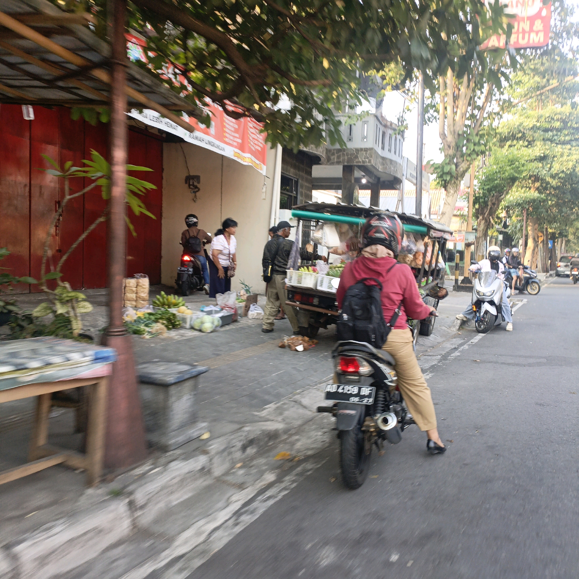 The image depicts a street scene in Salatiga, Indonesia, featuring a bustling sidewalk market and traffic. In the foreground, a person wearing a red hooded jacket and tan pants is seated on a motorcycle, their back to the camera, with a black backpack on. The motorcycle has a license plate reading "AD 4159 DF". To the left, a wooden table and bench are visible, with a potted plant.
Further down the sidewalk, a vendor is operating a food cart, with crates of fruits and vegetables displayed. Several people are interacting with the vendor and browsing the goods. Two women, one in a white shirt and the other in a patterned shirt, are standing near the cart. A man in a dark jacket and hat is also present. A person on a motorcycle with a black helmet is stopped near the cart.
In the background, more motorcycles are present, indicating typical street traffic. Buildings line the street, one with a red facade and another under construction or renovation with stone elements and balconies. Trees and foliage are abundant, suggesting a green urban environment. The time of day appears to be late afternoon, given the lighting conditions and the long shadows. The overall atmosphere is one of everyday commerce and activity in a city setting.