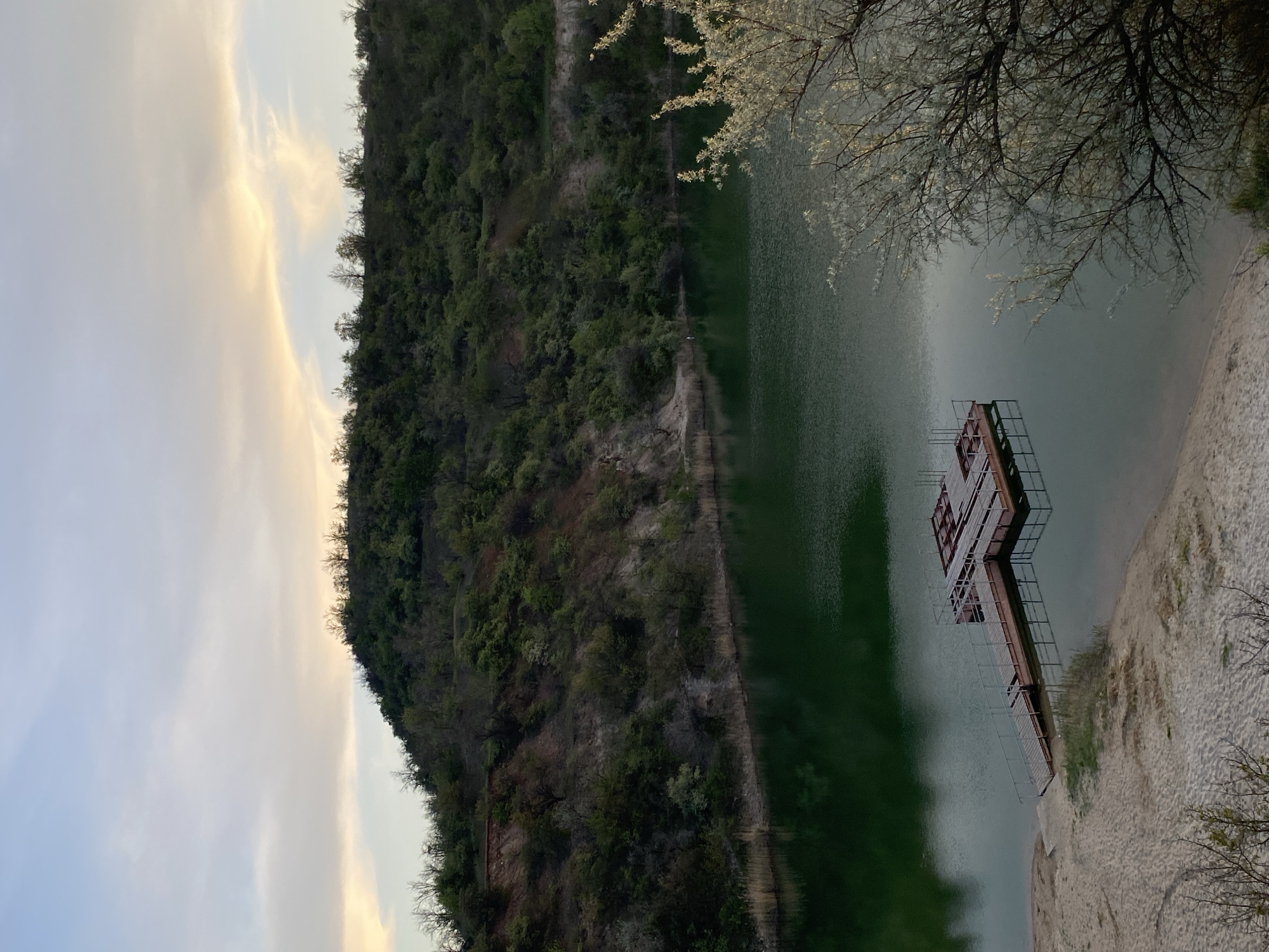 A landscape photograph depicts a lake, likely in an unknown city, set against the backdrop of a tree-covered hill under a sky with light-colored clouds. A wooden dock extends into the lake from a sandy shoreline on the right side of the frame. The hill, covered in dense vegetation, runs along the left side, providing a contrast to the lighter sky.